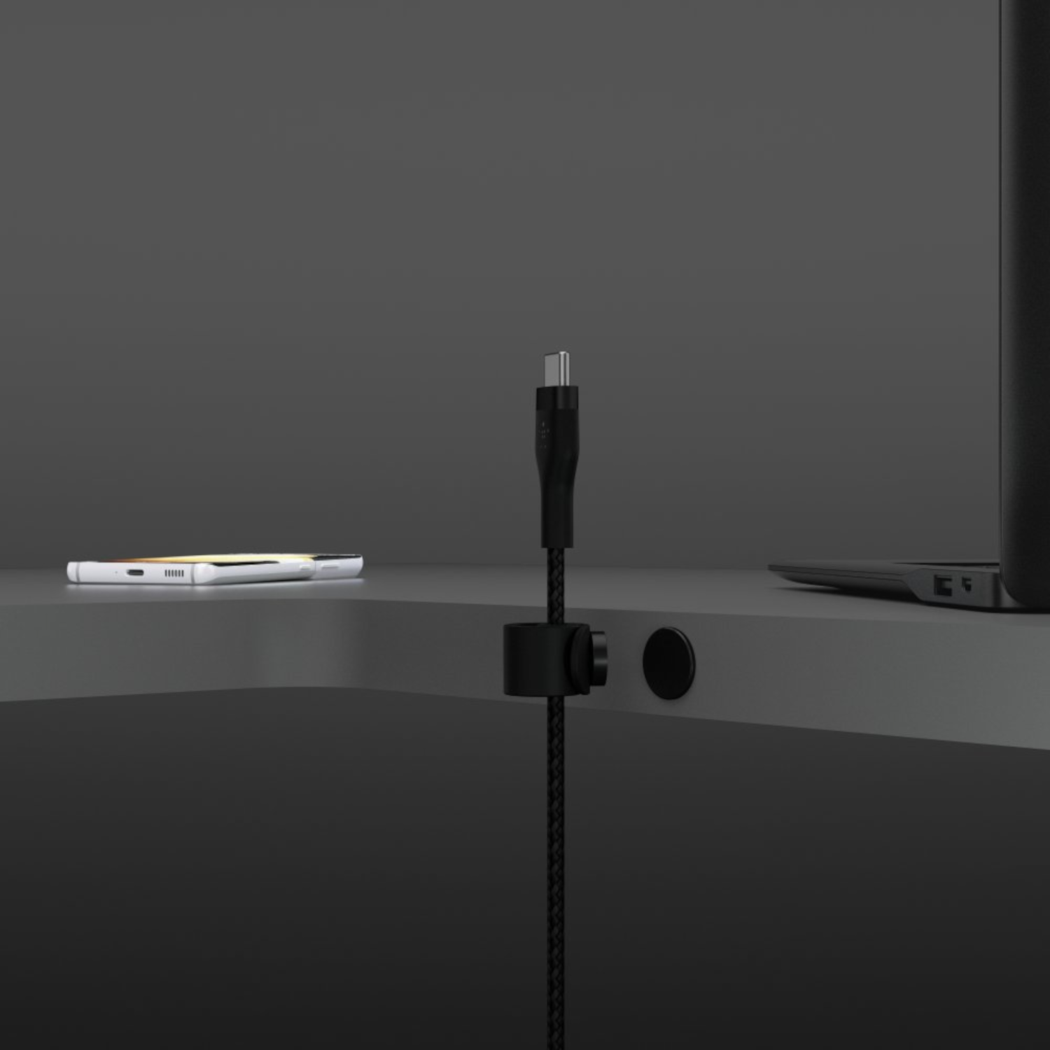 Un câble de chargement noir est connecté à un ordinateur portable. Un téléphone est à gauche. Le câble est tenu par un clip noir. Le fond est gris.