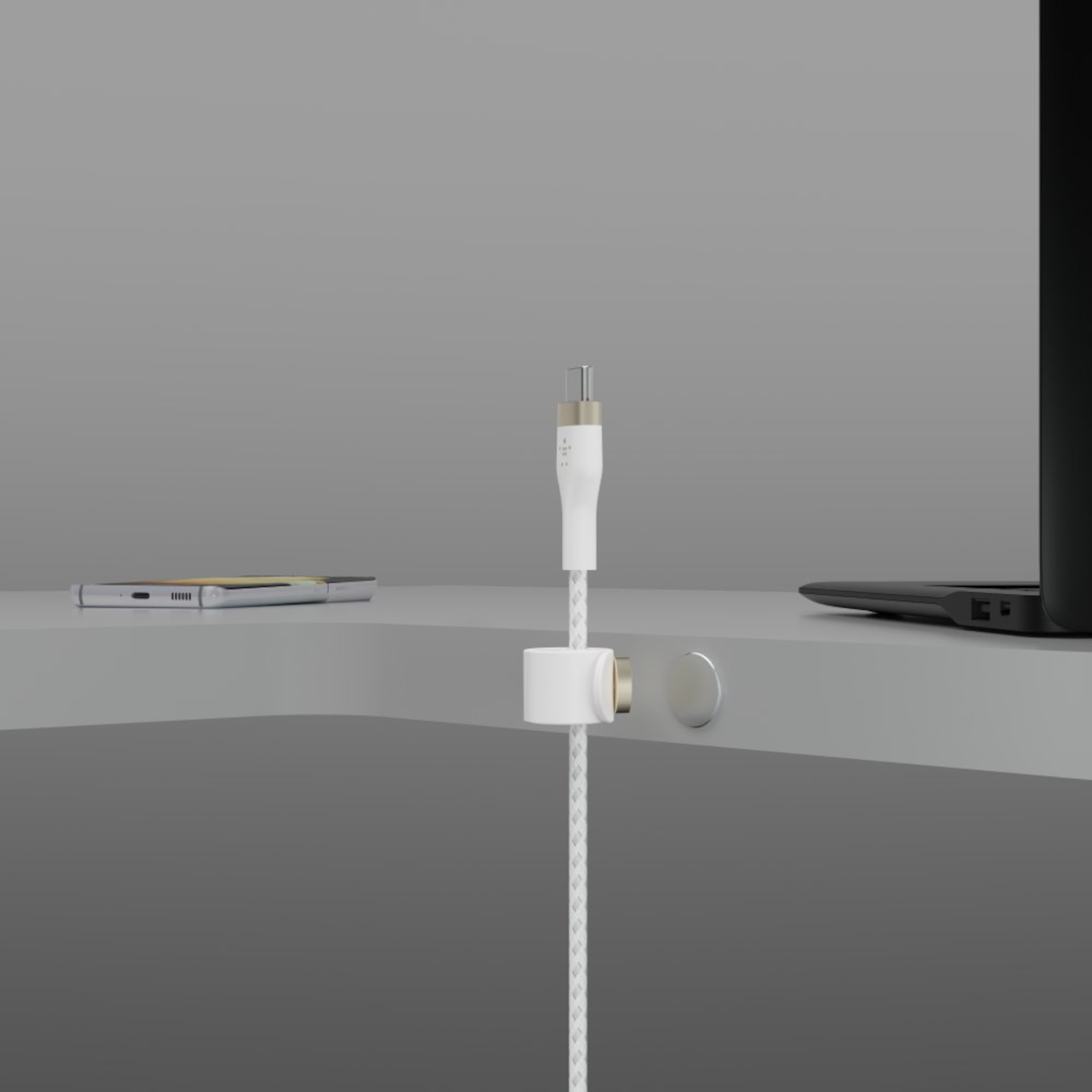 Un câble de recharge blanc avec un support magnétique. Un téléphone et un ordinateur portable sont visibles sur une surface grise, fond gris.
