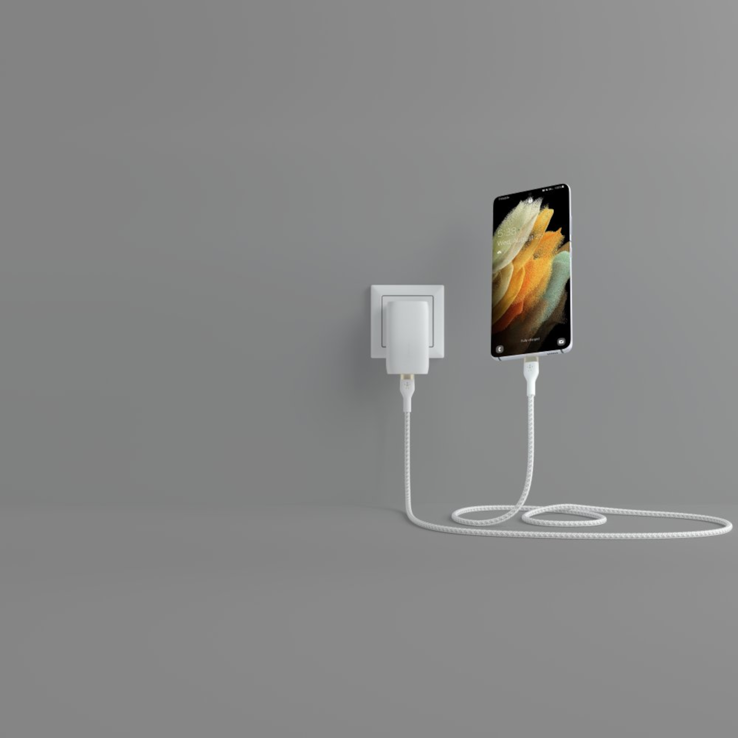 Un smartphone est connecté à un chargeur branché sur une prise murale. L'écran du téléphone est allumé, affichant la date.
