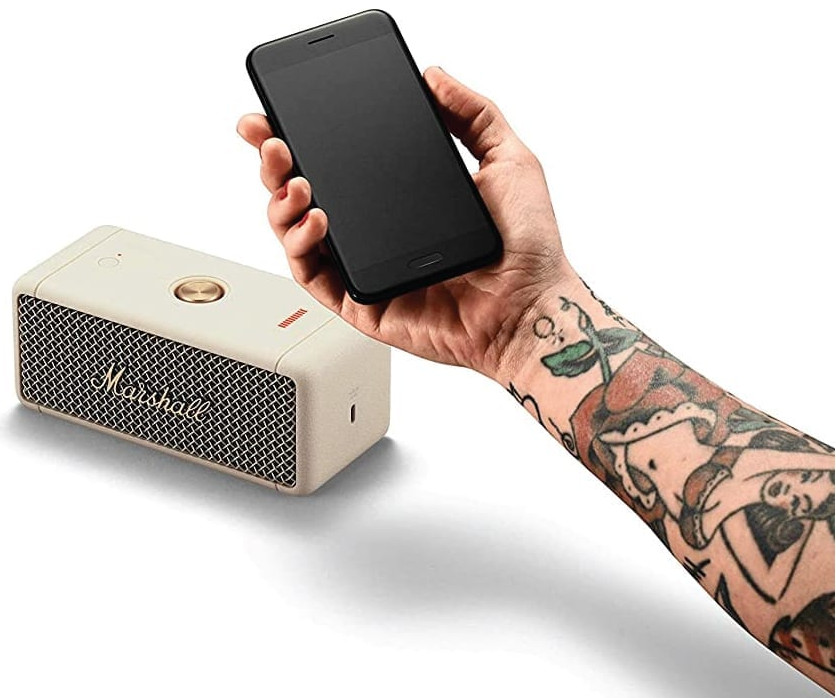 Een persoon met tatoeages die een telefoon vasthoudt bij een beige speaker.