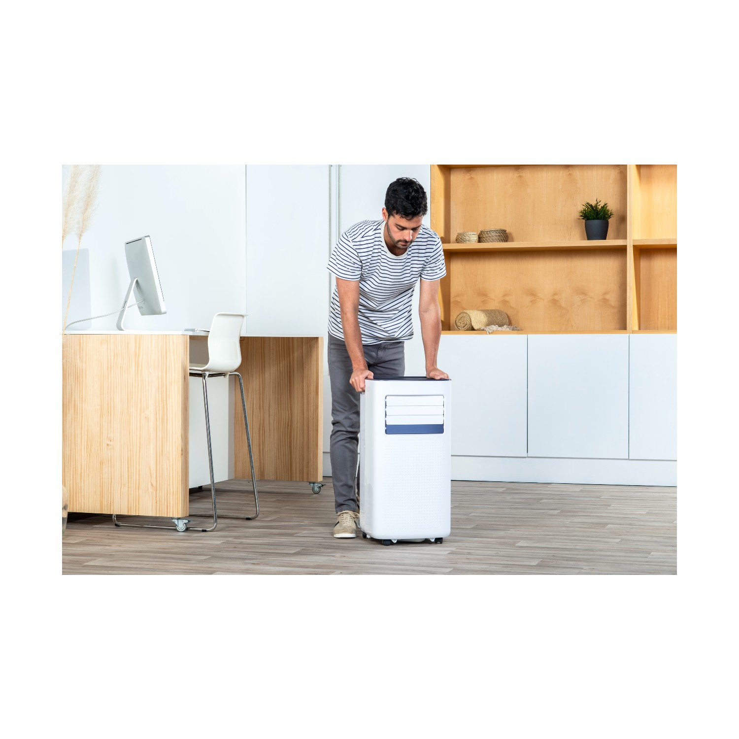 Hombre rodando un aire acondicionado portátil blanco. La habitación tiene gabinetes de madera y pared blanca.