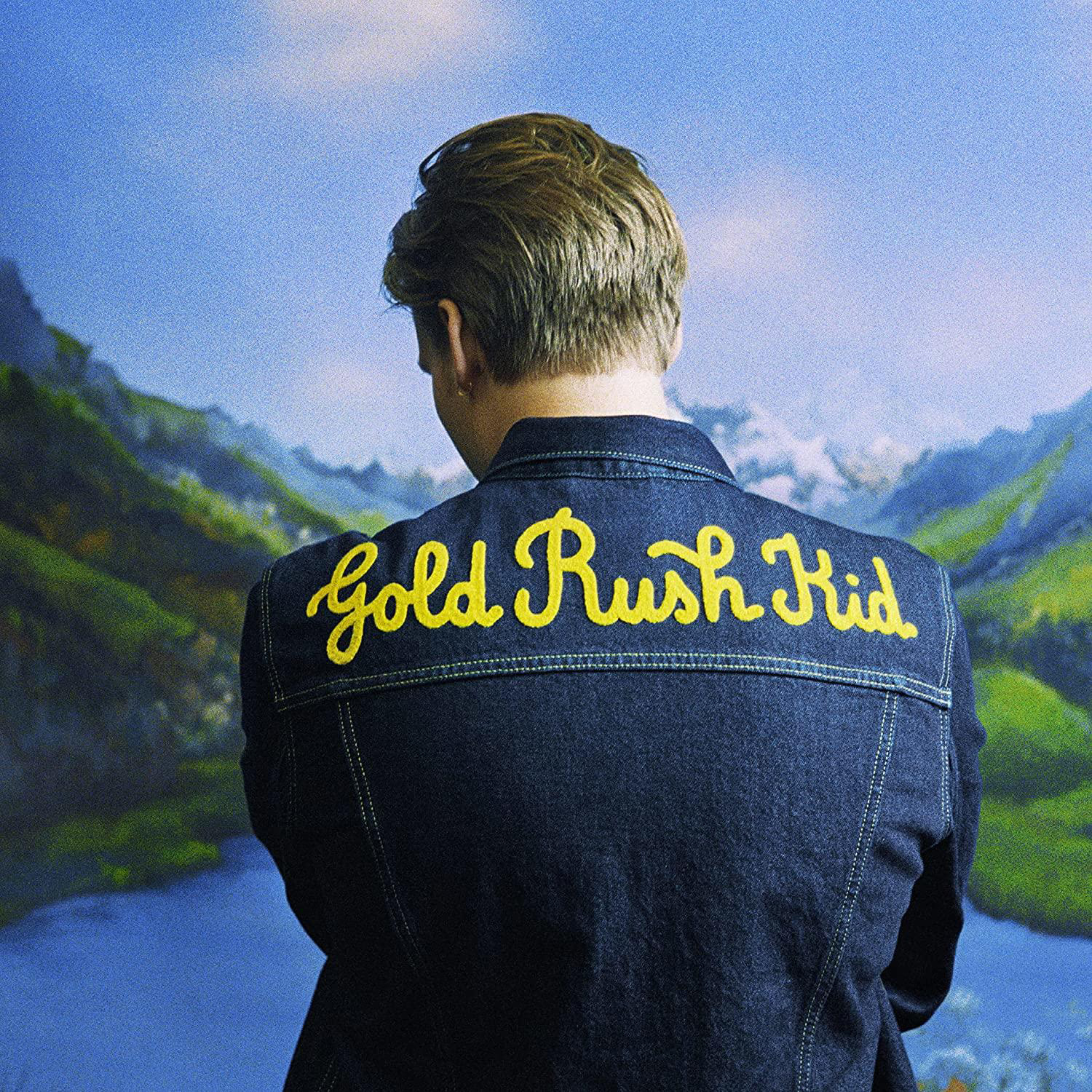 Mann in Jeansjacke, Rücken zur Kamera, gelber Text: Gold Rush Kid, Bergkulisse.