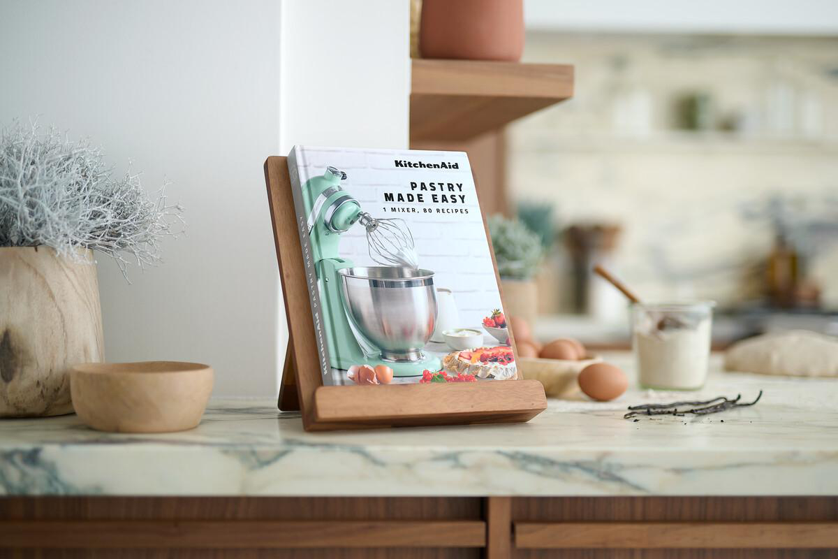 Ein Kochbuch auf einem Holzständer, ein KitchenAid-Mixer, Eier, Zutaten und eine Schüssel auf der Theke.