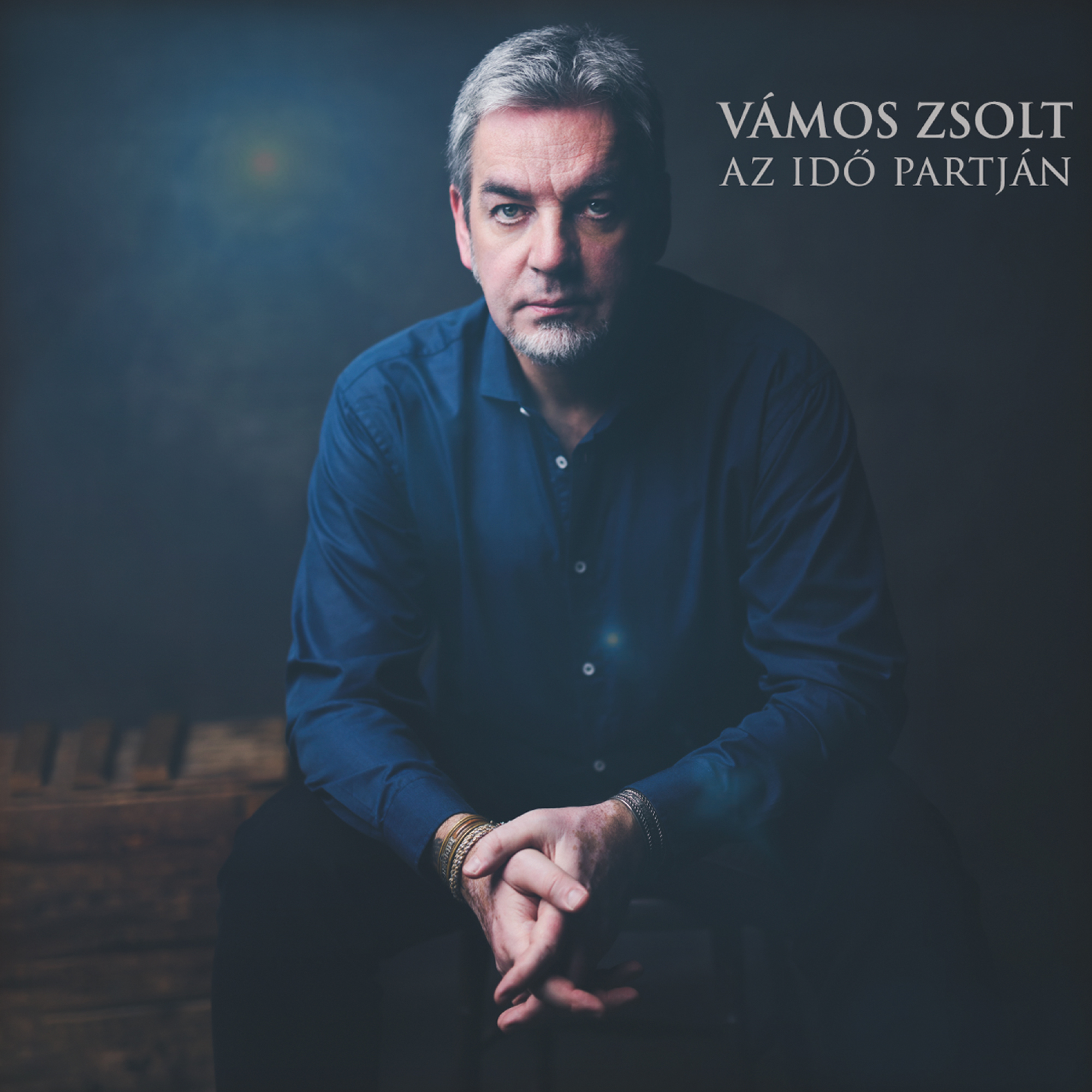 Egy szürke hajú, kék inges férfi, összefont kézzel ül. A háttér sötét.
