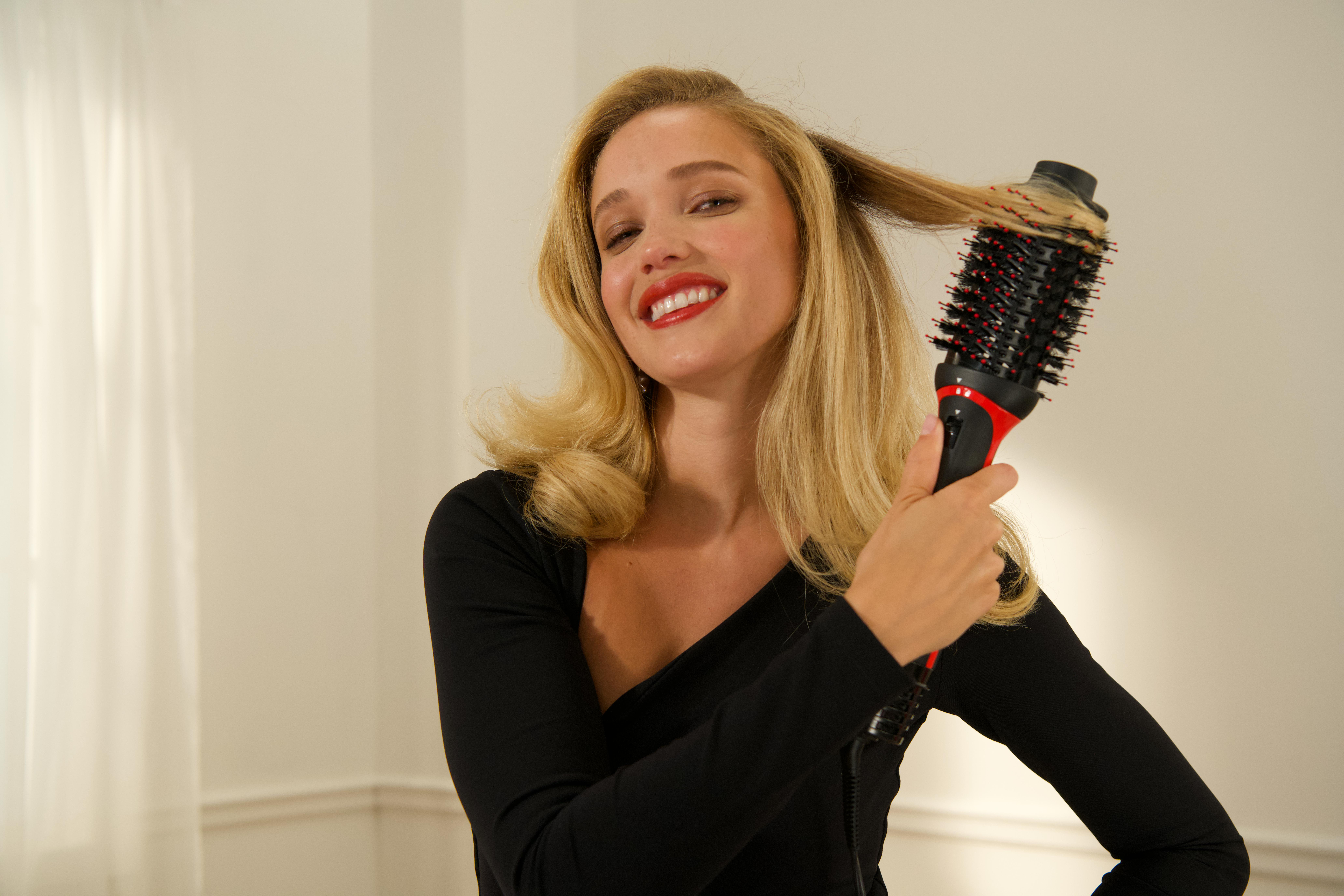 Blonde Frau, die sich mit einer Rundbürste die Haare bürstet, lächelt, schwarzes Oberteil.