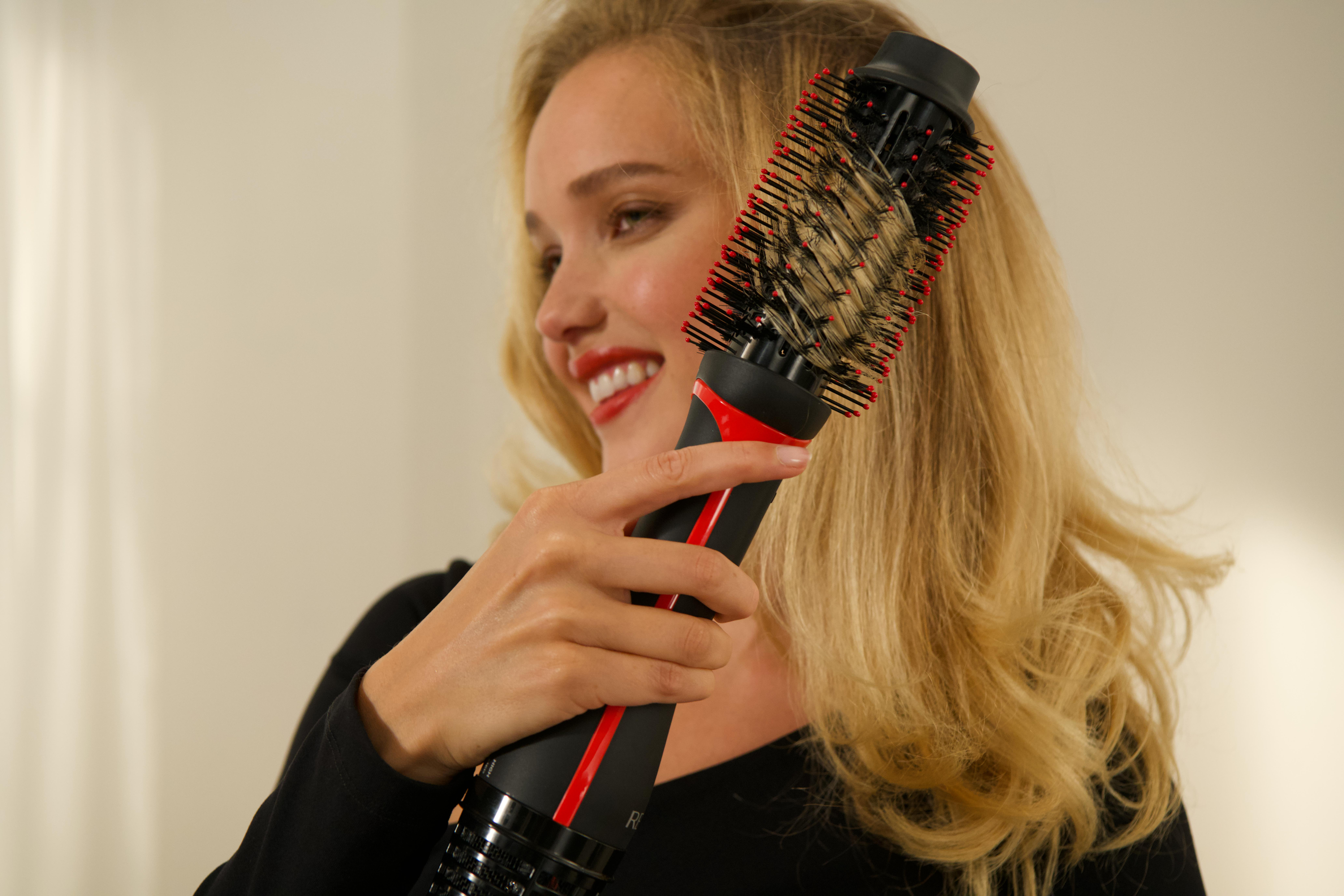 Frau benutzt eine Haartrocknerbürste, lächelt, blondes Haar, rote und schwarze Bürste.