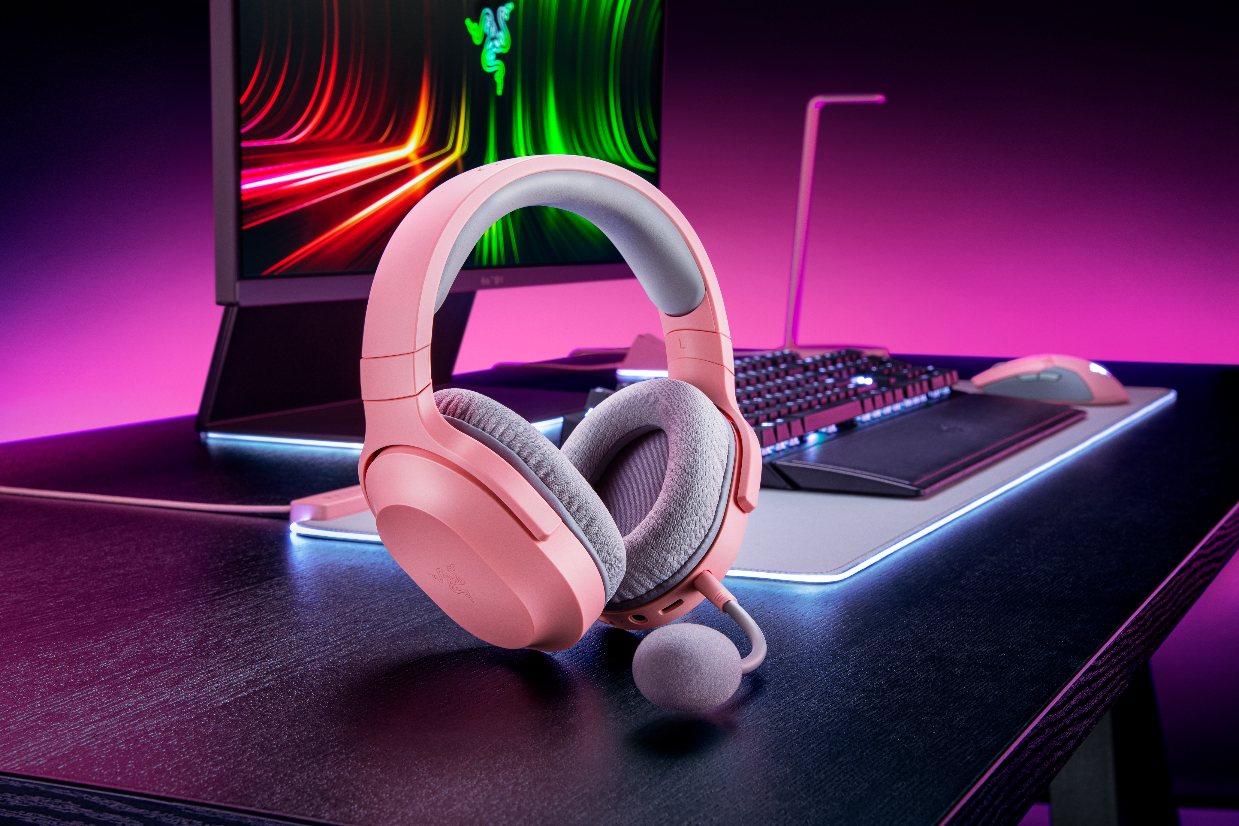 Cuffie da gioco rosa, microfono, tastiera e mouse su una scrivania scura con sfondo rosa e viola.
