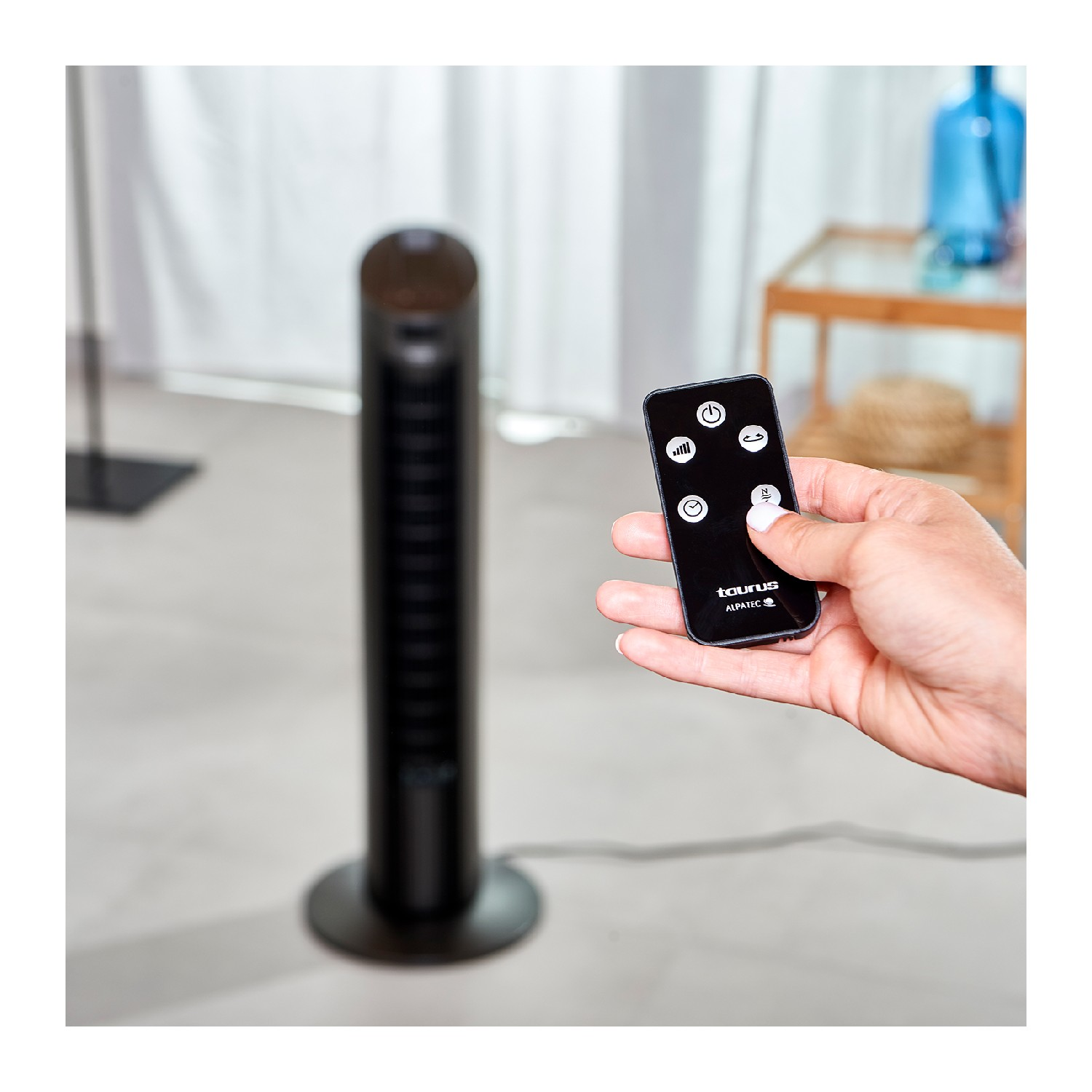 Persona sosteniendo un mando a distancia cerca de un ventilador de torre negro. El ventilador está sobre una superficie blanca.