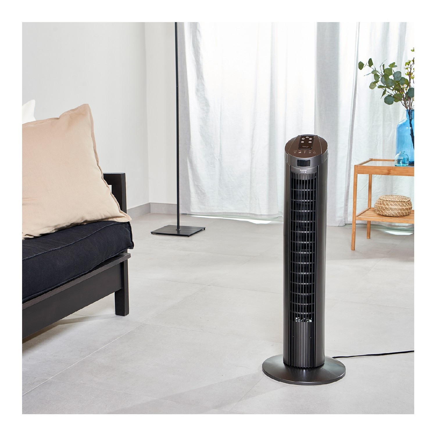 Un ventilador de torre negro está en una sala de estar. Hay un sofá y una ventana en el fondo.