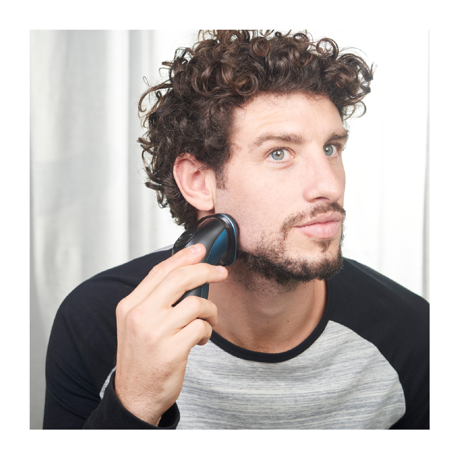 Un hombre afeitándose la barba con una afeitadora eléctrica, mirando a la cámara.
