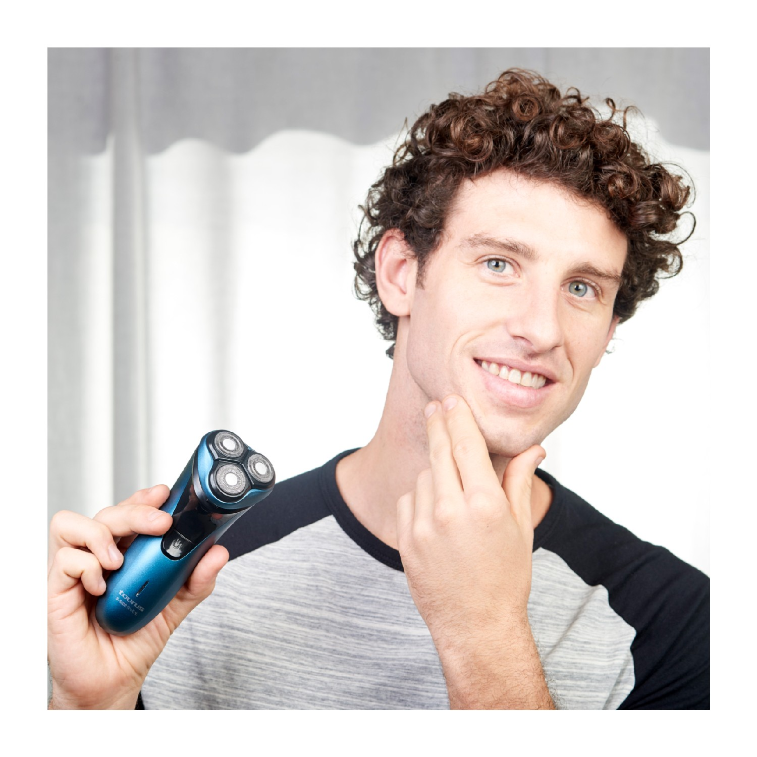 Hombre sonriendo, sosteniendo una afeitadora eléctrica azul. Se está tocando la cara.