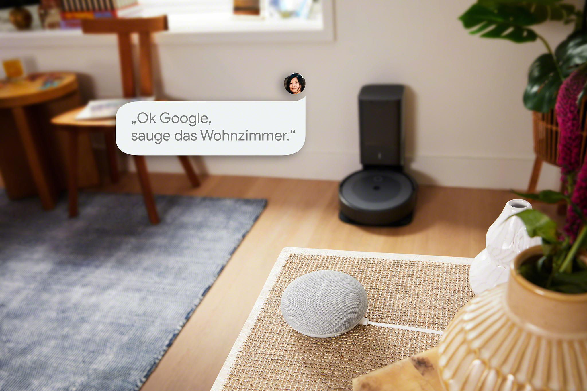 Ein Smart-Home-Setup mit einem Google Nest Mini-Lautsprecher und einem Roboterstaubsauger. Eine Sprechblase sagt: Ok Google, sauge das Wohnzimmer.
