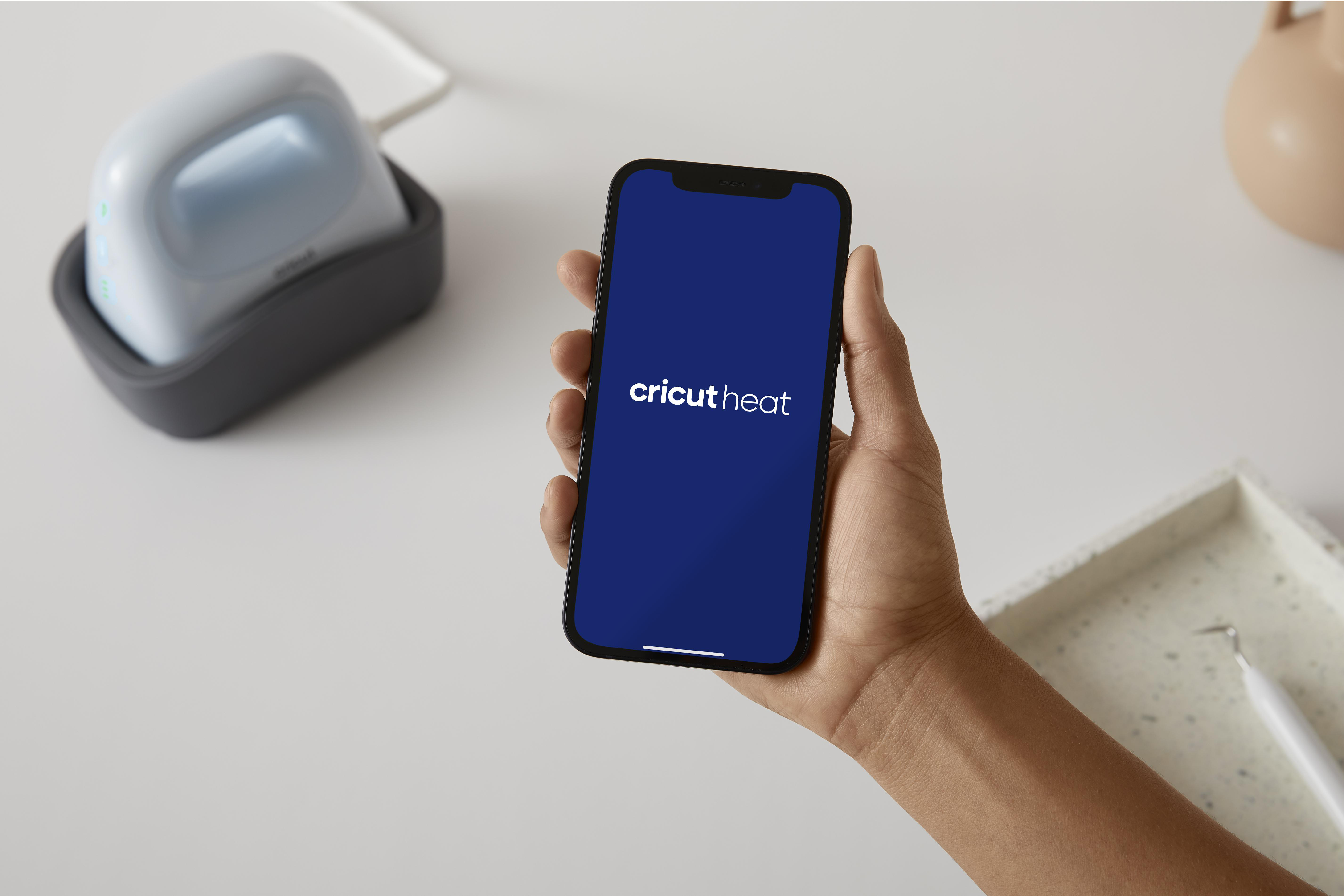 Eine Hand, die ein Smartphone mit dem Logo 'cricut heat' auf dem Bildschirm hält. Links befindet sich eine blaue Thermopresse.