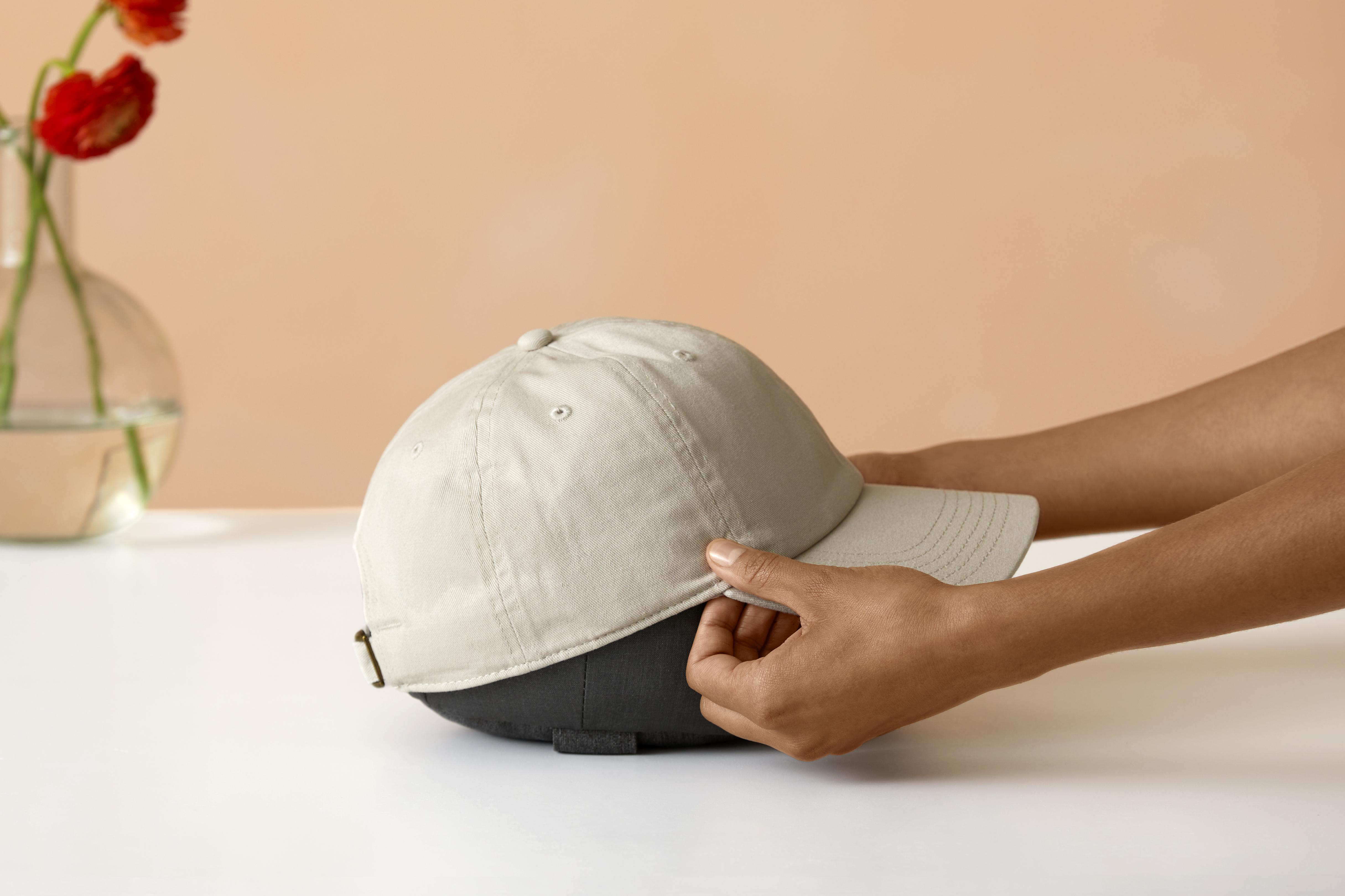 Eine beige Kappe wird von Händen auf einen dunkelgrauen Hutständer gelegt. Weicher, neutraler Hintergrund.