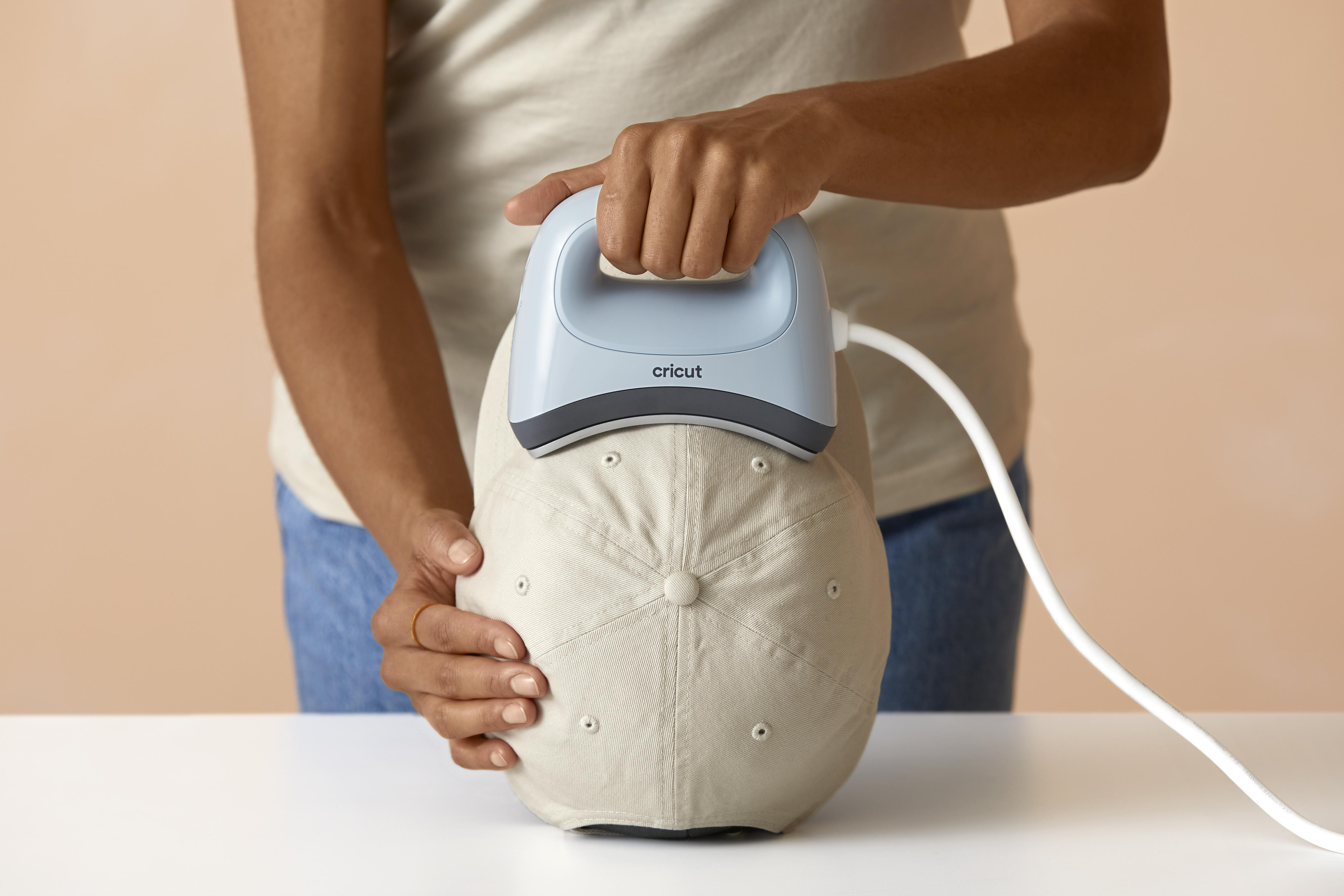 Person verwendet Cricut EasyPress auf einer Baseballkappe. Hellblaue Presse, beige Kappe, heller Hintergrund.