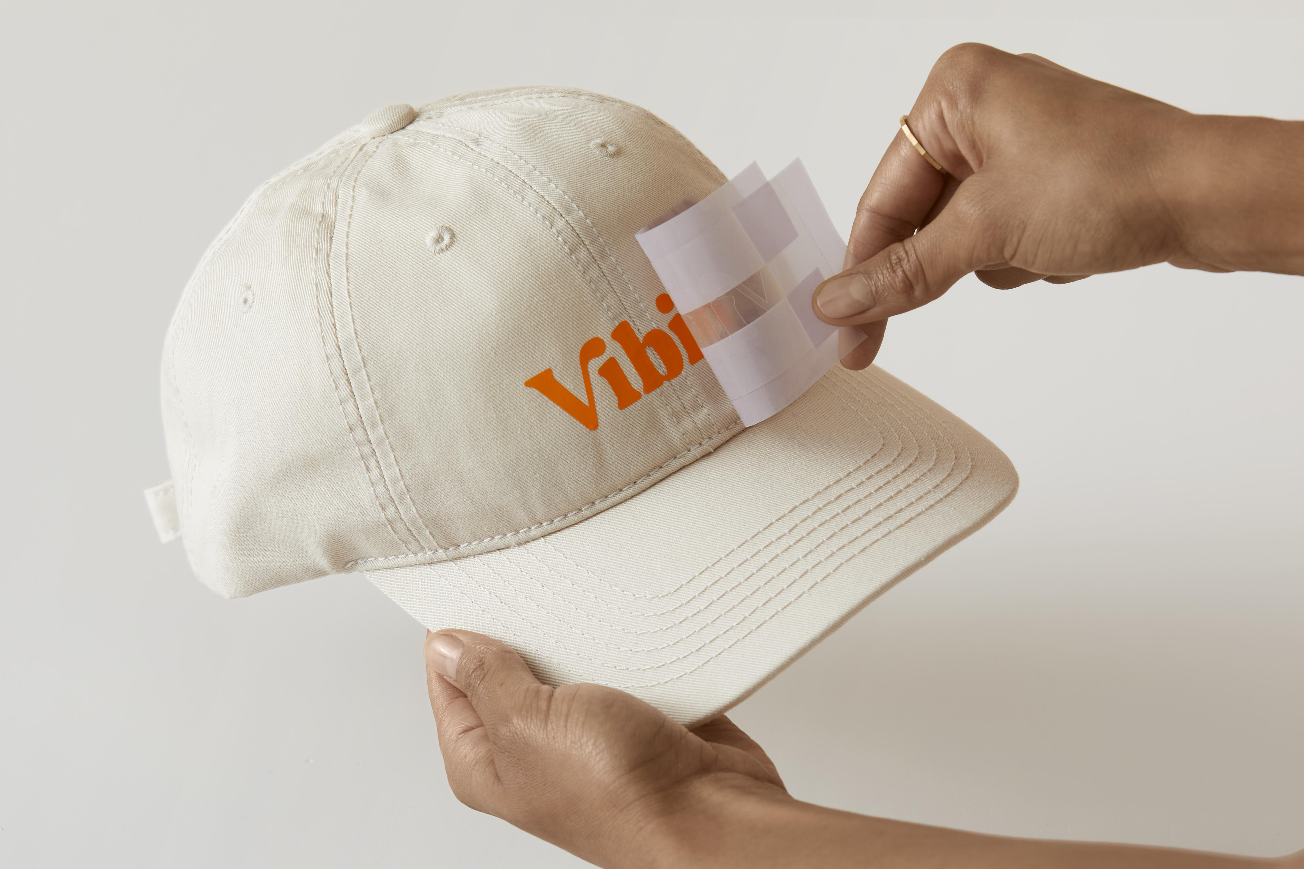 Eine Hand, die einen weißen Hut mit Logo hält, und einen Aufkleber anbringt. Das Logo ist orange.