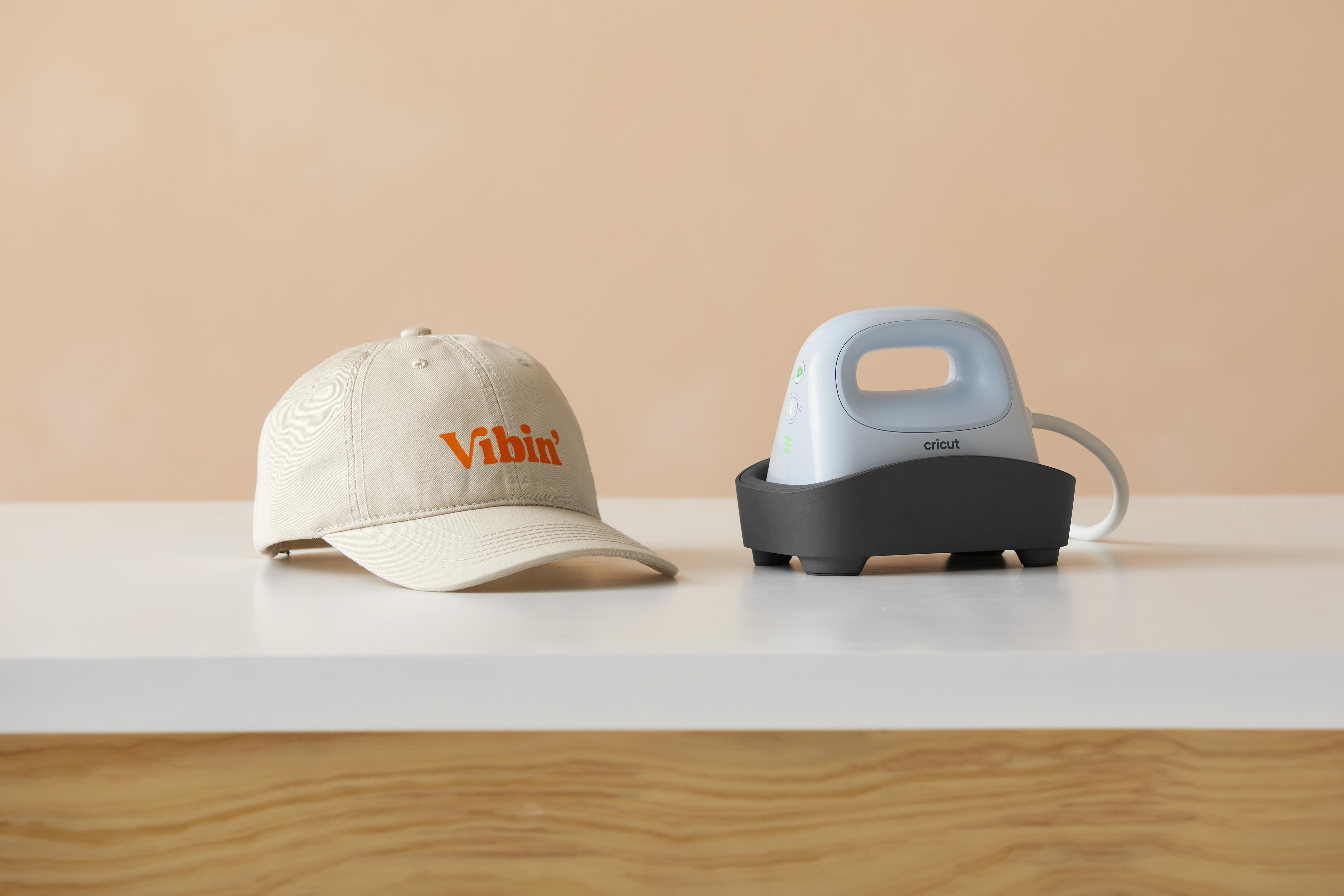 Eine weiße Baseballkappe mit orangefarbenem Text 'Vibin', neben einer kleinen blauen Cricut-Thermobresse.