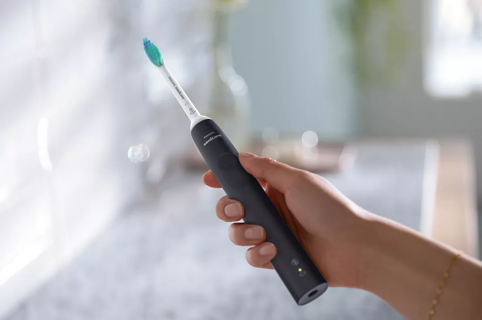 Une personne tenant une brosse à dents électrique noire, avec une tête de brosse bleu-vert, dans une salle de bain.