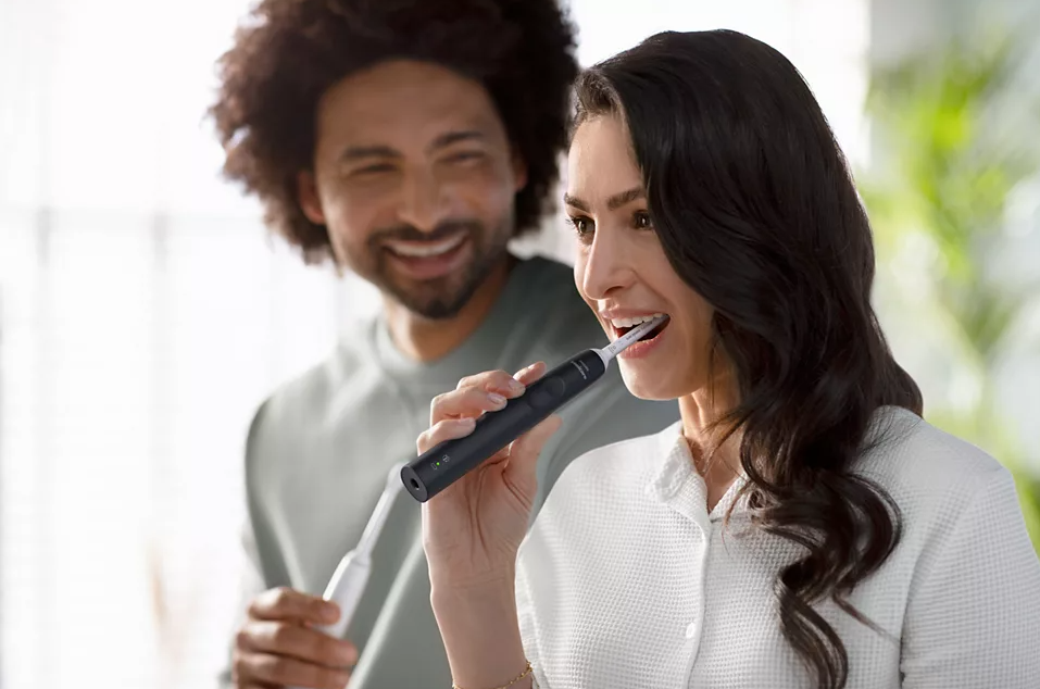 Un homme et une femme souriant, se brossant les dents avec des brosses à dents électriques. Brosse à dents noire au centre.