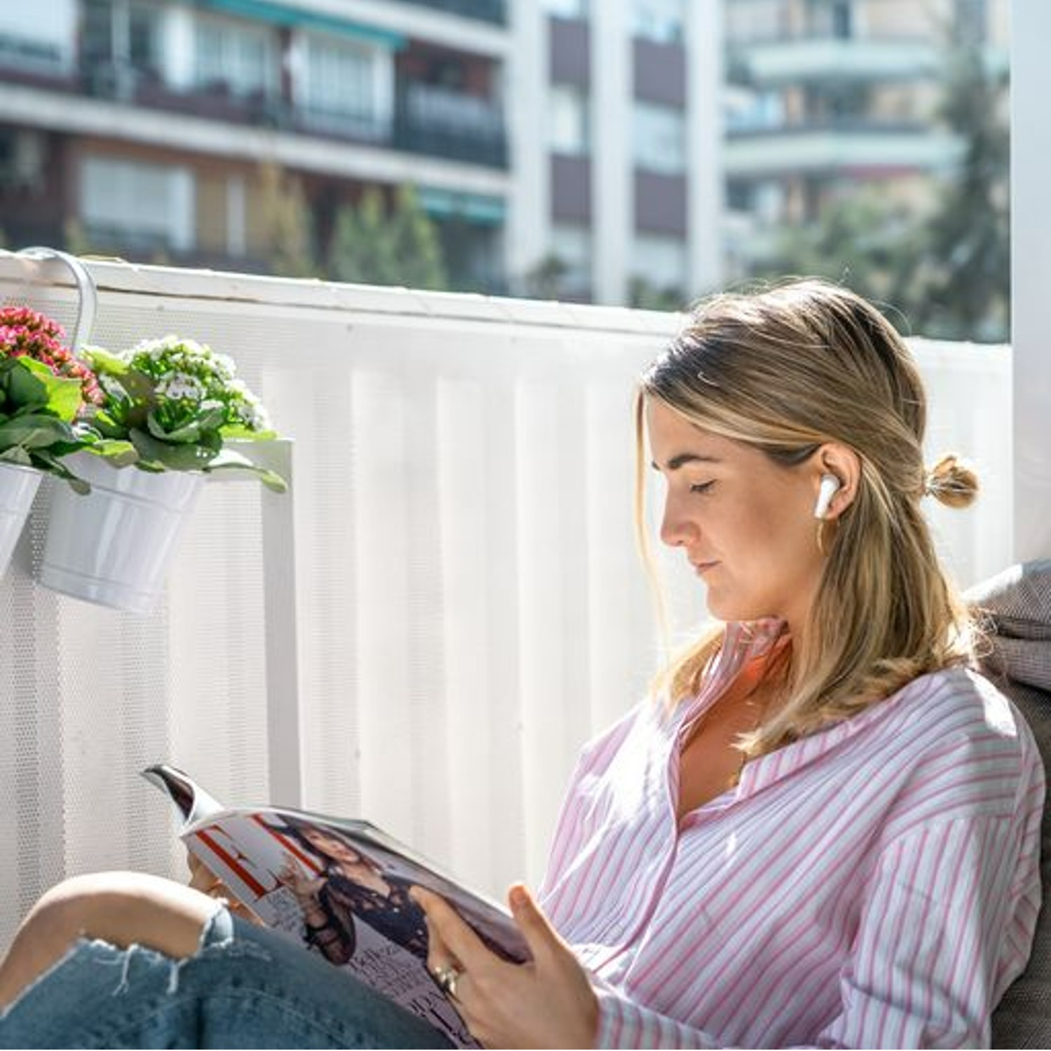 Una mujer en un balcón lee una revista, usa auriculares y una camisa a rayas rosa.