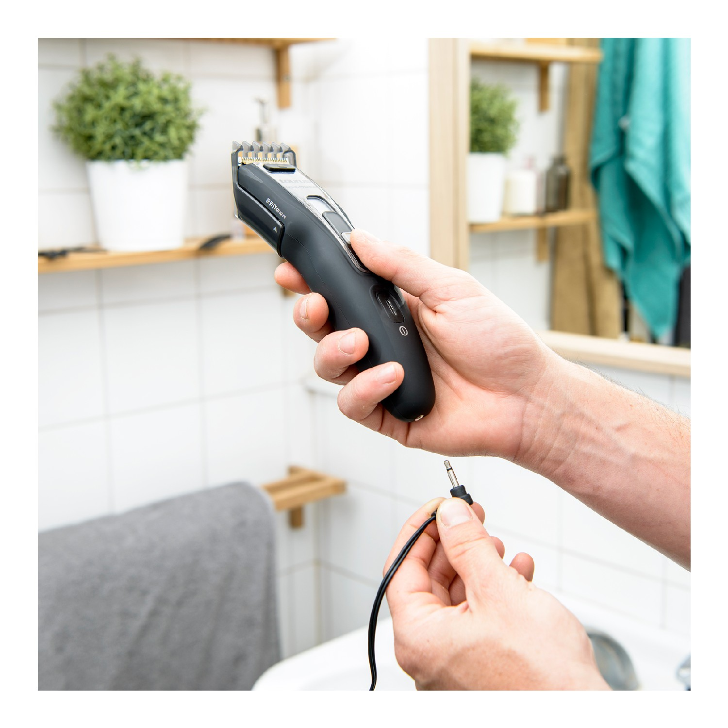 Una persona sosteniendo una recortadora negra con un cable negro, en un baño, cerca de un espejo.