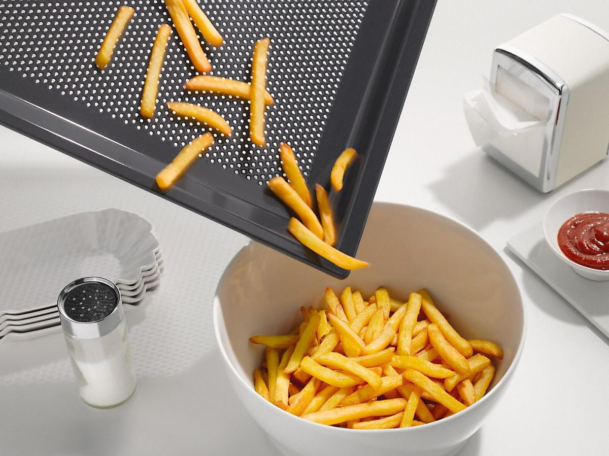 Pommes frites werden von einem Tablett in eine weiße Schüssel gegossen, Salz, Ketchup und ein Serviettenhalter daneben.