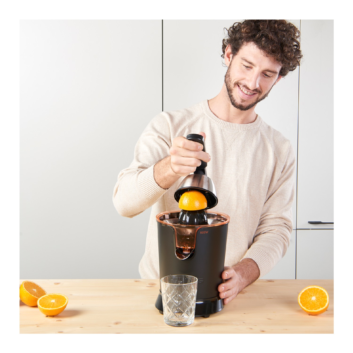 Un hombre exprimiendo una naranja con un exprimidor. El exprimidor es negro y cobrizo. Hay un vaso y mitades de naranja.