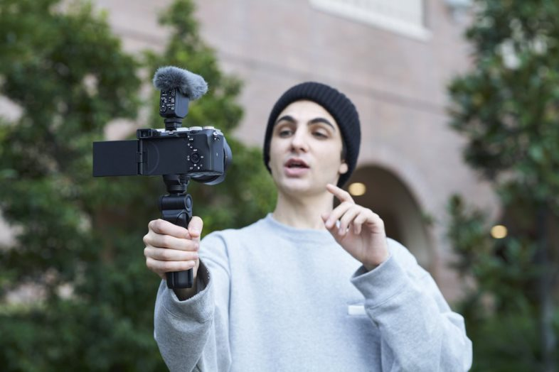 Homme tenant une caméra avec un microphone. Il porte un bonnet.