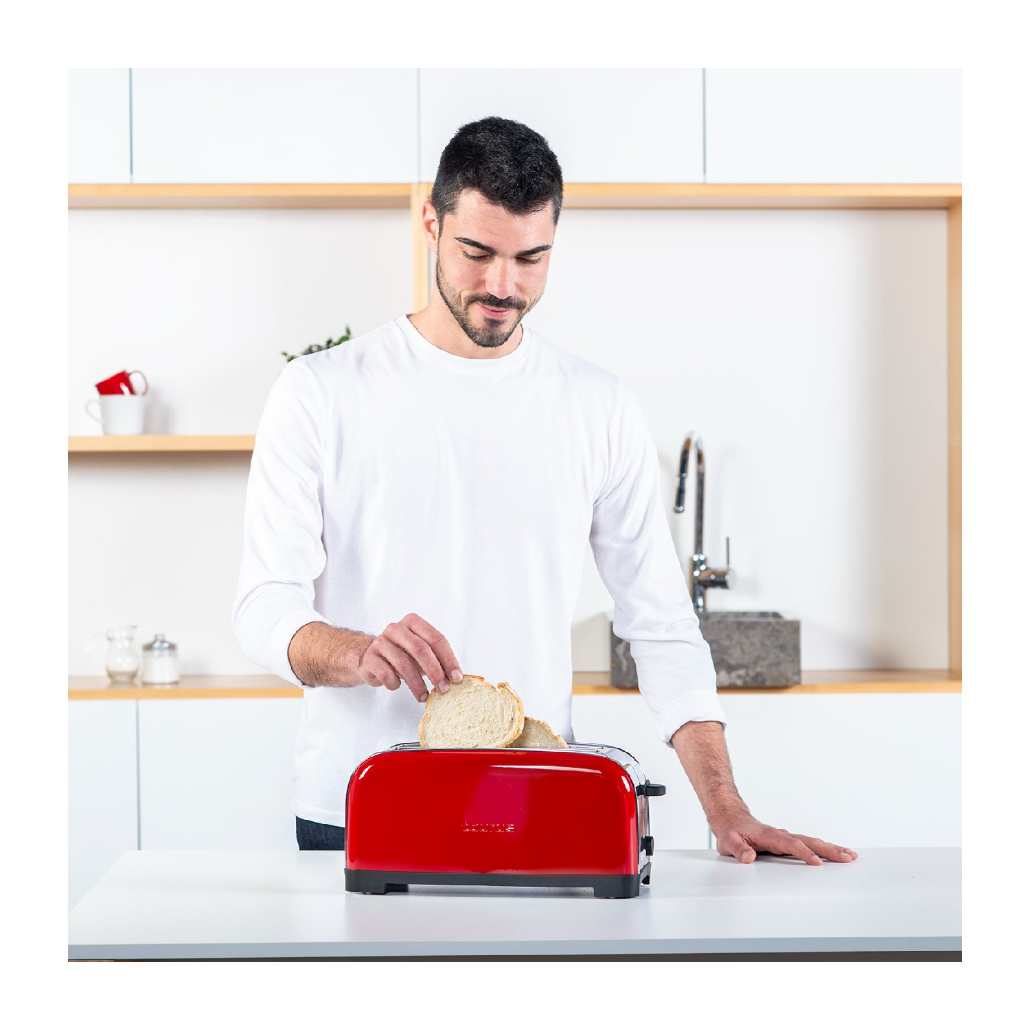 Hombre poniendo pan en una tostadora roja. Cocina blanca. El hombre lleva una camisa blanca de manga larga.