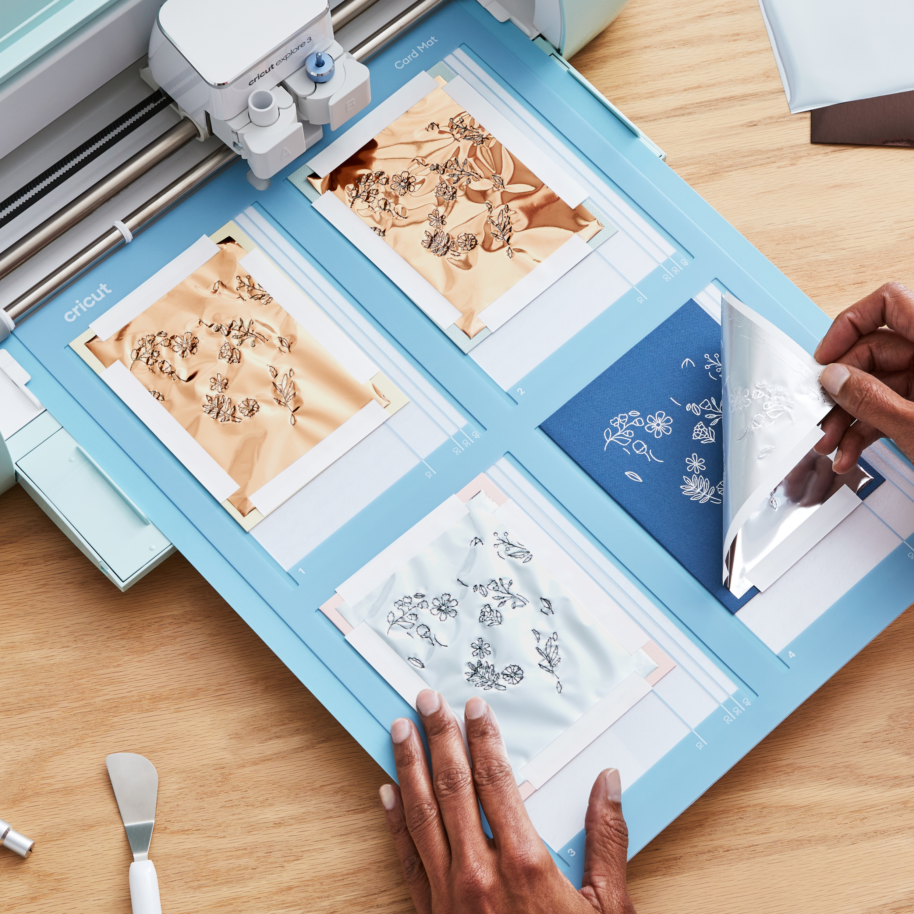 Eine Cricut-Maschine schneidet Folie auf einer blauen Matte. Hände ziehen Folie von einer blauen Karte mit Blumendesign ab.