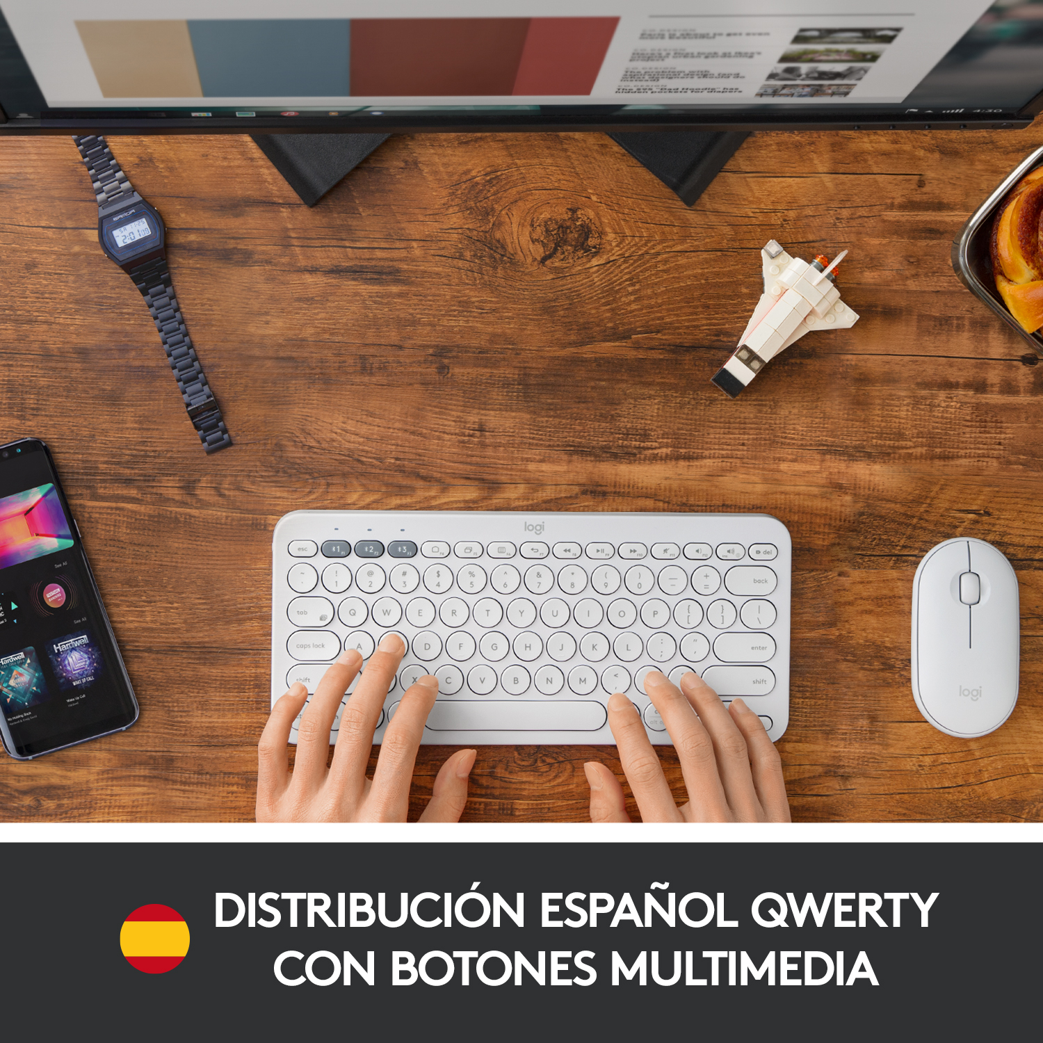 Manos tecleando en un teclado blanco con diseño QWERTY español, con otros dispositivos y un reloj.