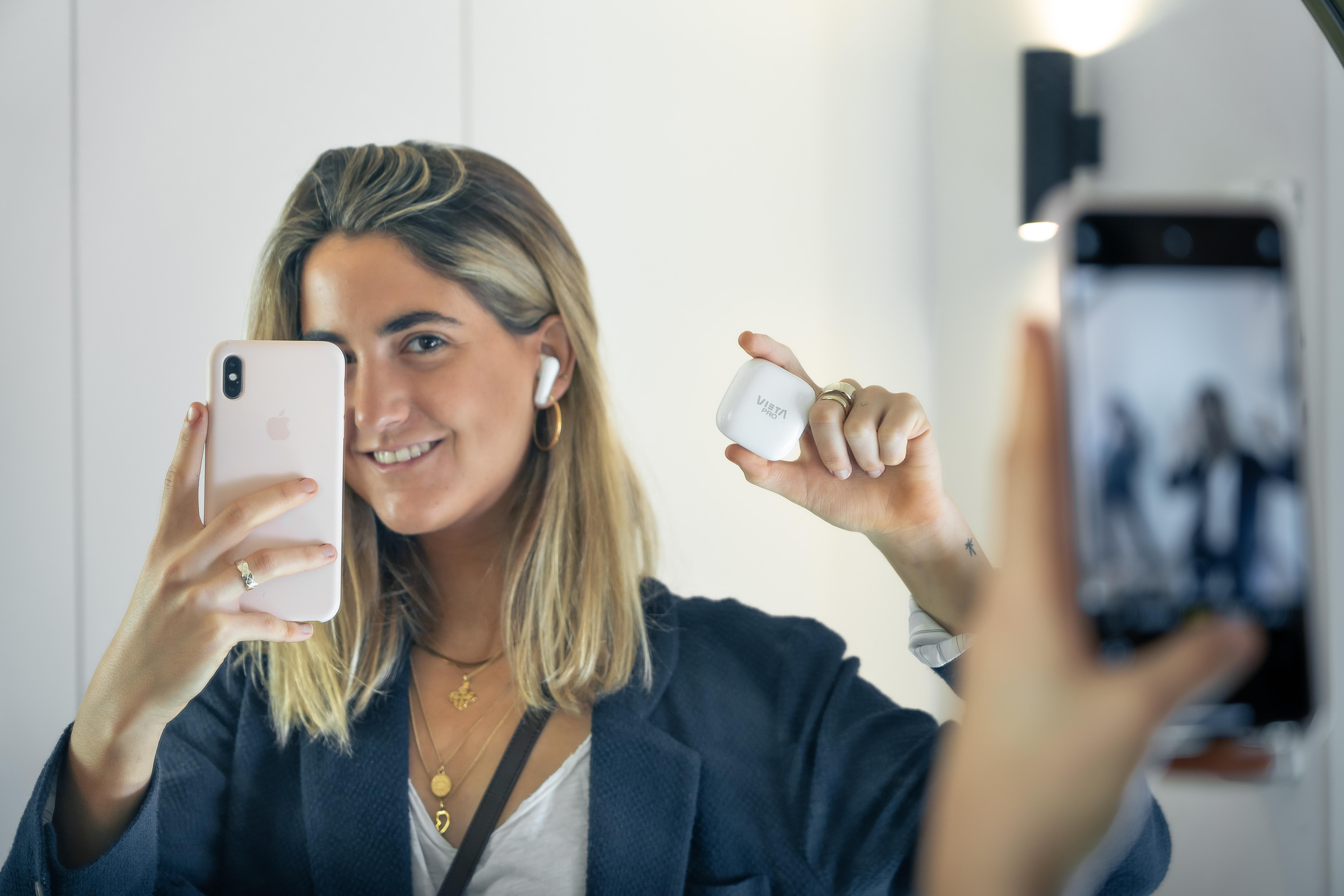 Frau hält ein Telefon und Ohrhörer, macht ein Selfie mit einem quadratischen Ohrhörer-Etui.