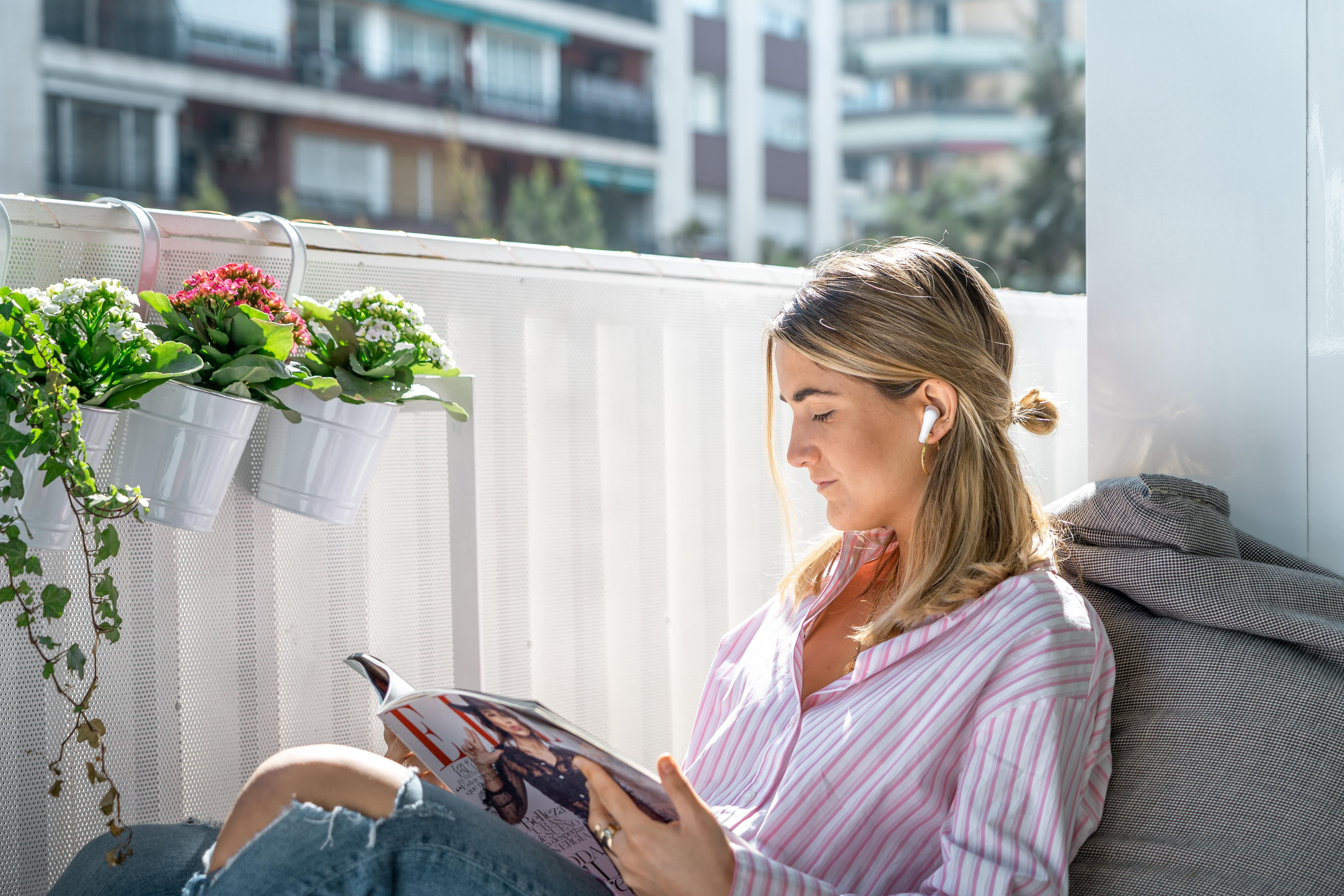 Eine Frau liest auf einem Balkon eine Zeitschrift und trägt weiße Ohrhörer.