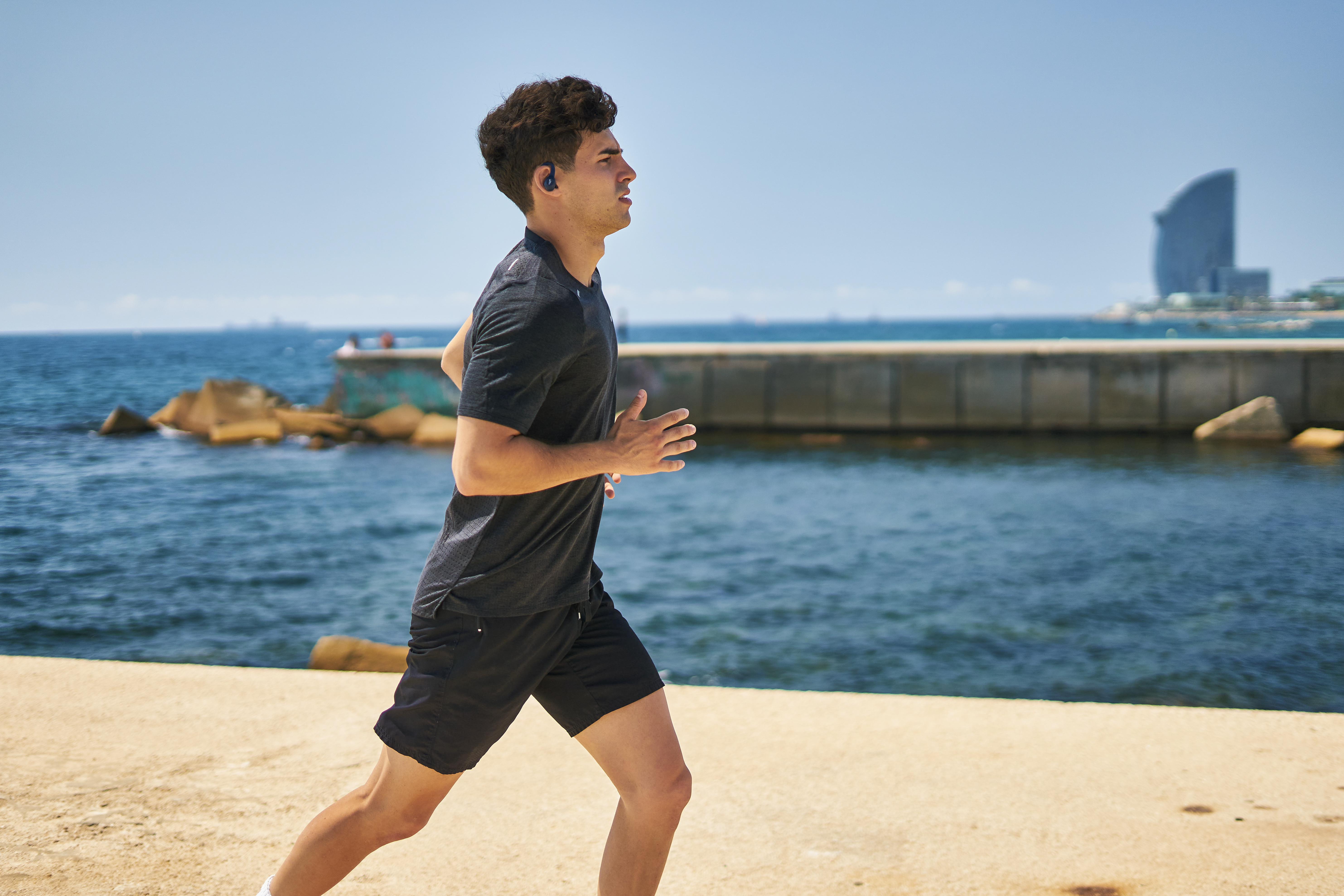 Mann rennt am Strand, schwarzes Shirt und Shorts, Gebäude im Hintergrund.