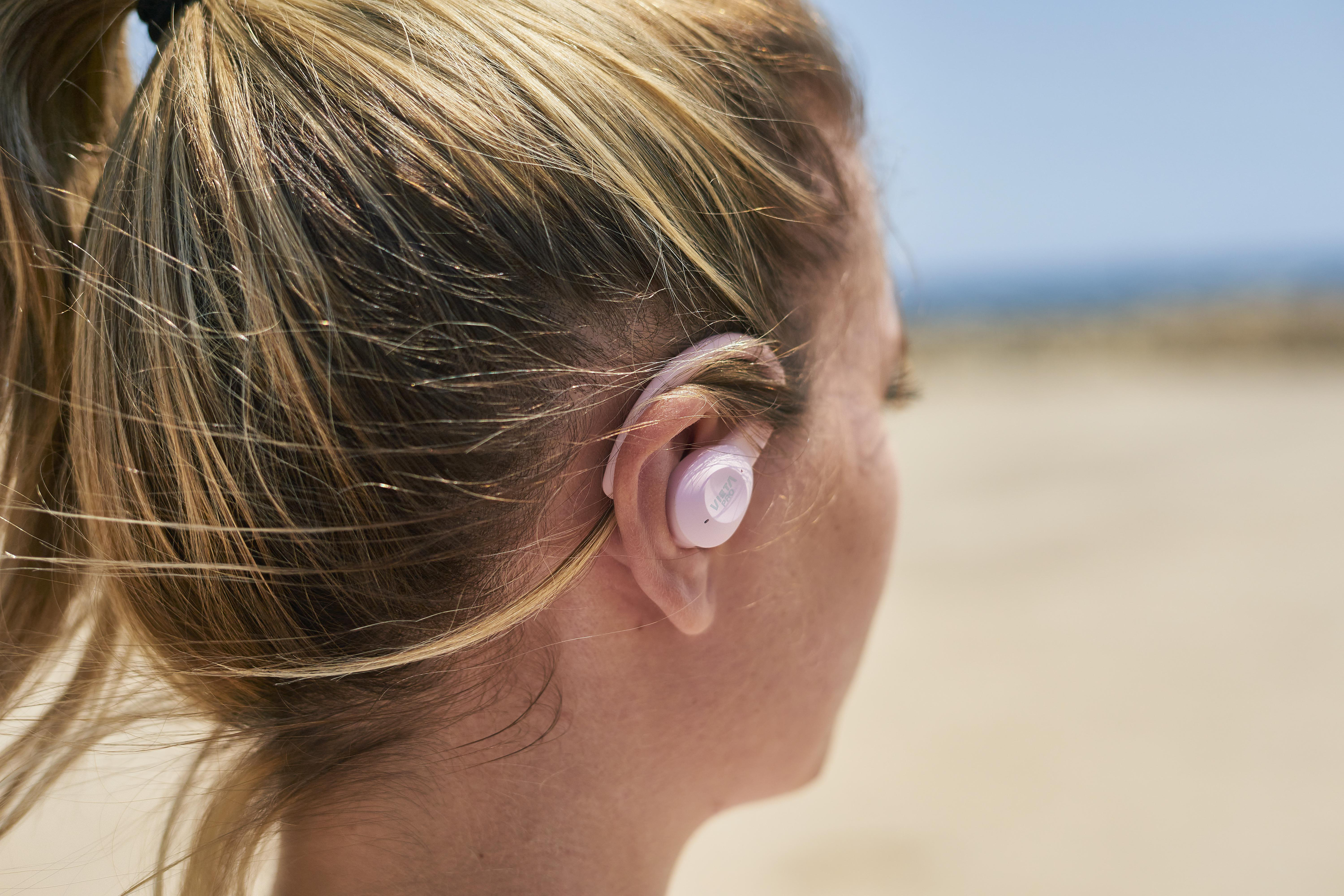 Ohr einer Frau mit pinkem Ohrhörer, Strand im Hintergrund.