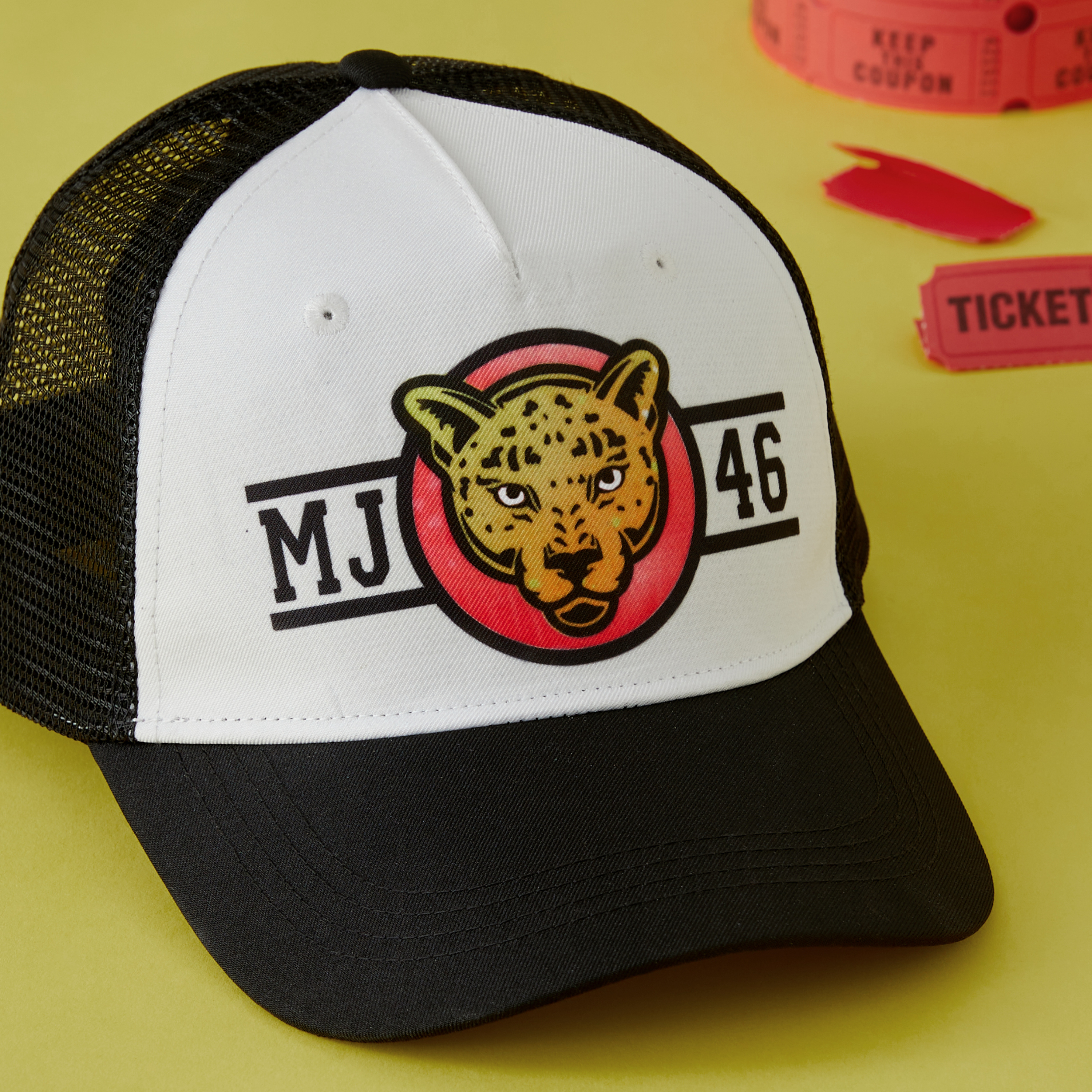 Eine schwarz-weiße Trucker-Mütze mit einem Leoparden-Logo und roten Tickets auf gelbem Untergrund.