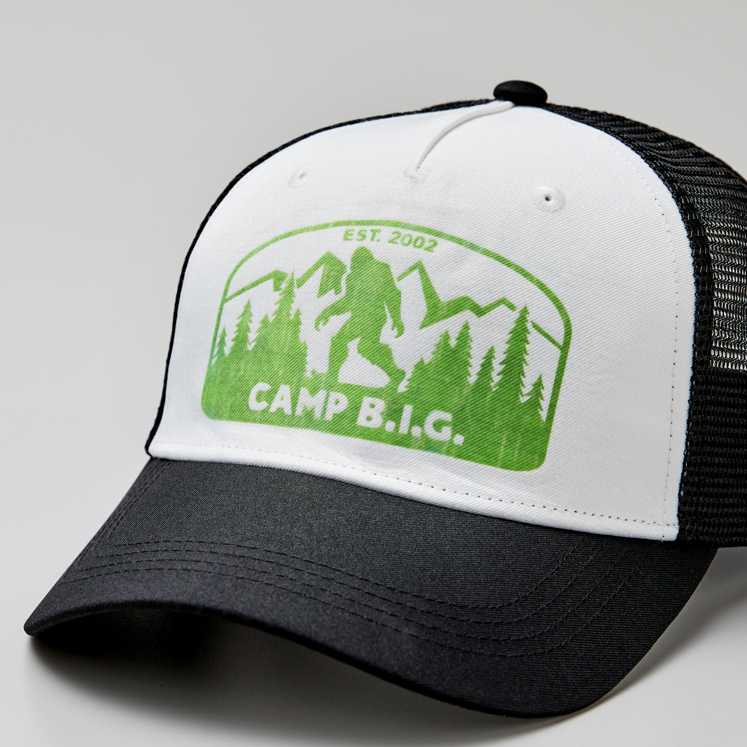 Eine weiß-schwarze Kappe mit grünem Logo eines Bigfoot, Bäumen und Bergen mit dem Text 'CAMP B.I.G.'