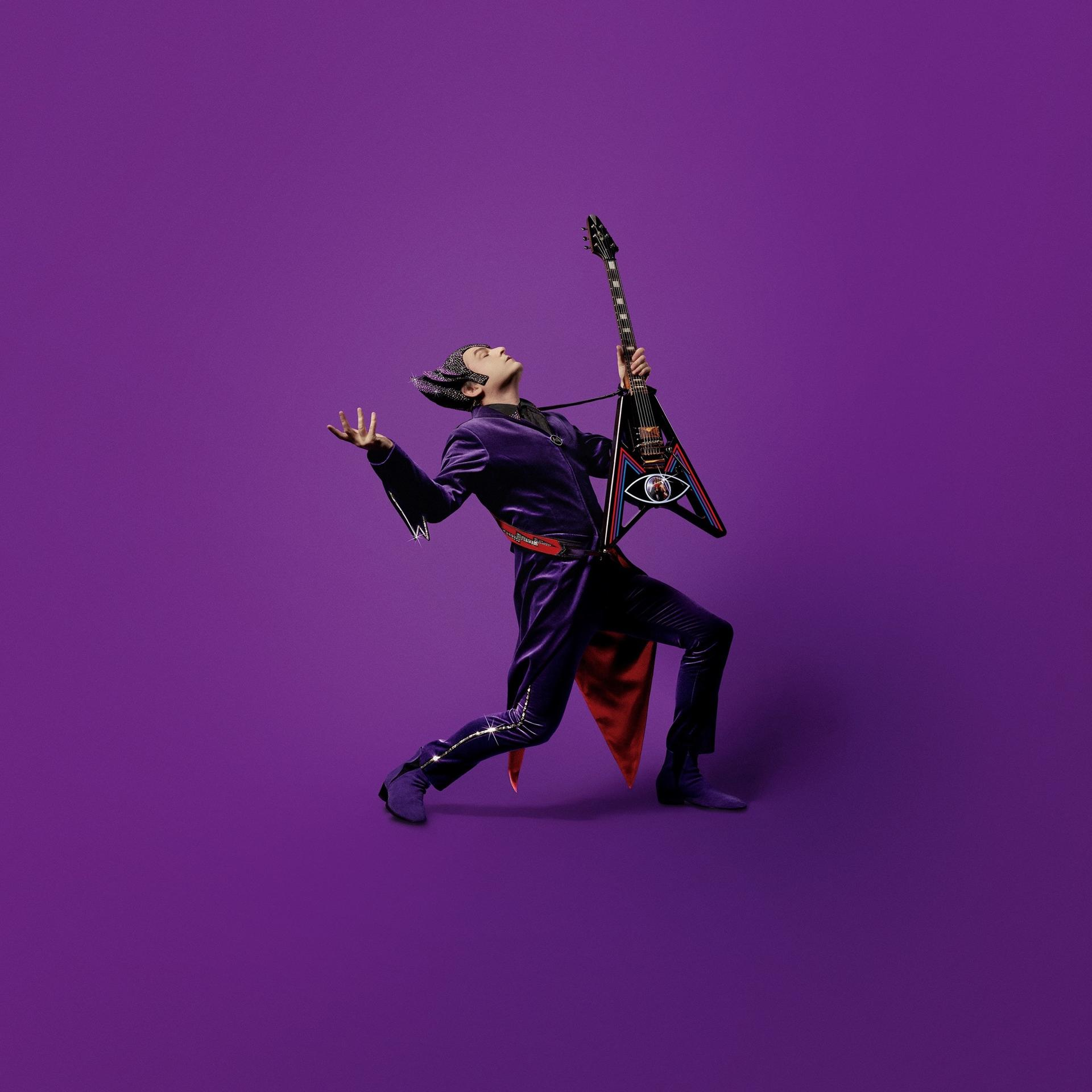 A man in a purple suit playing a custom guitar. He is on a purple background.