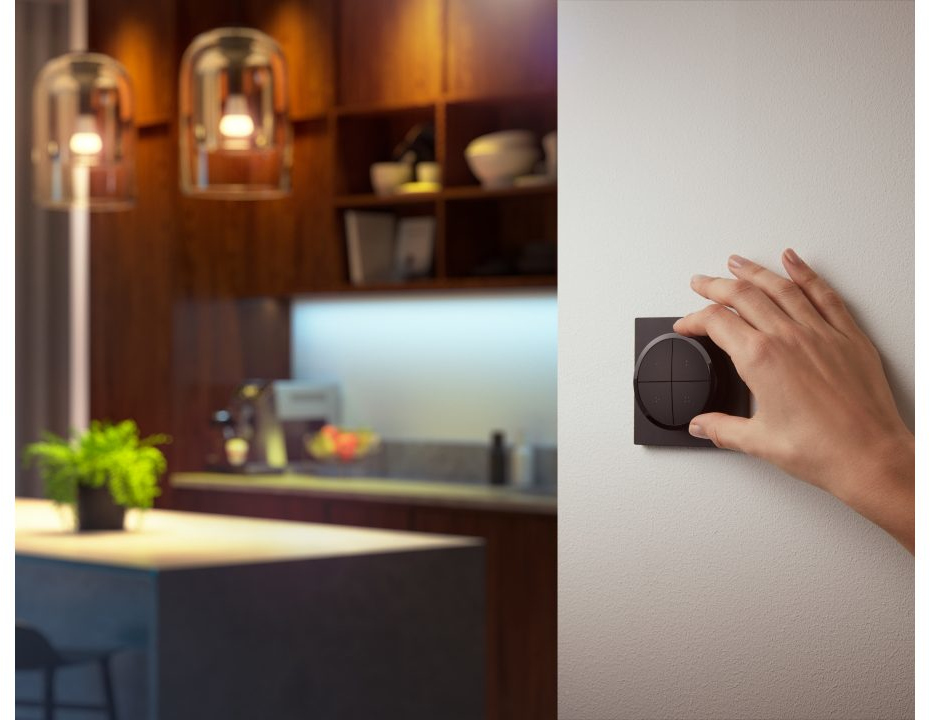 Hand touching a smart home control panel on a wall, kitchen and lights in the background.