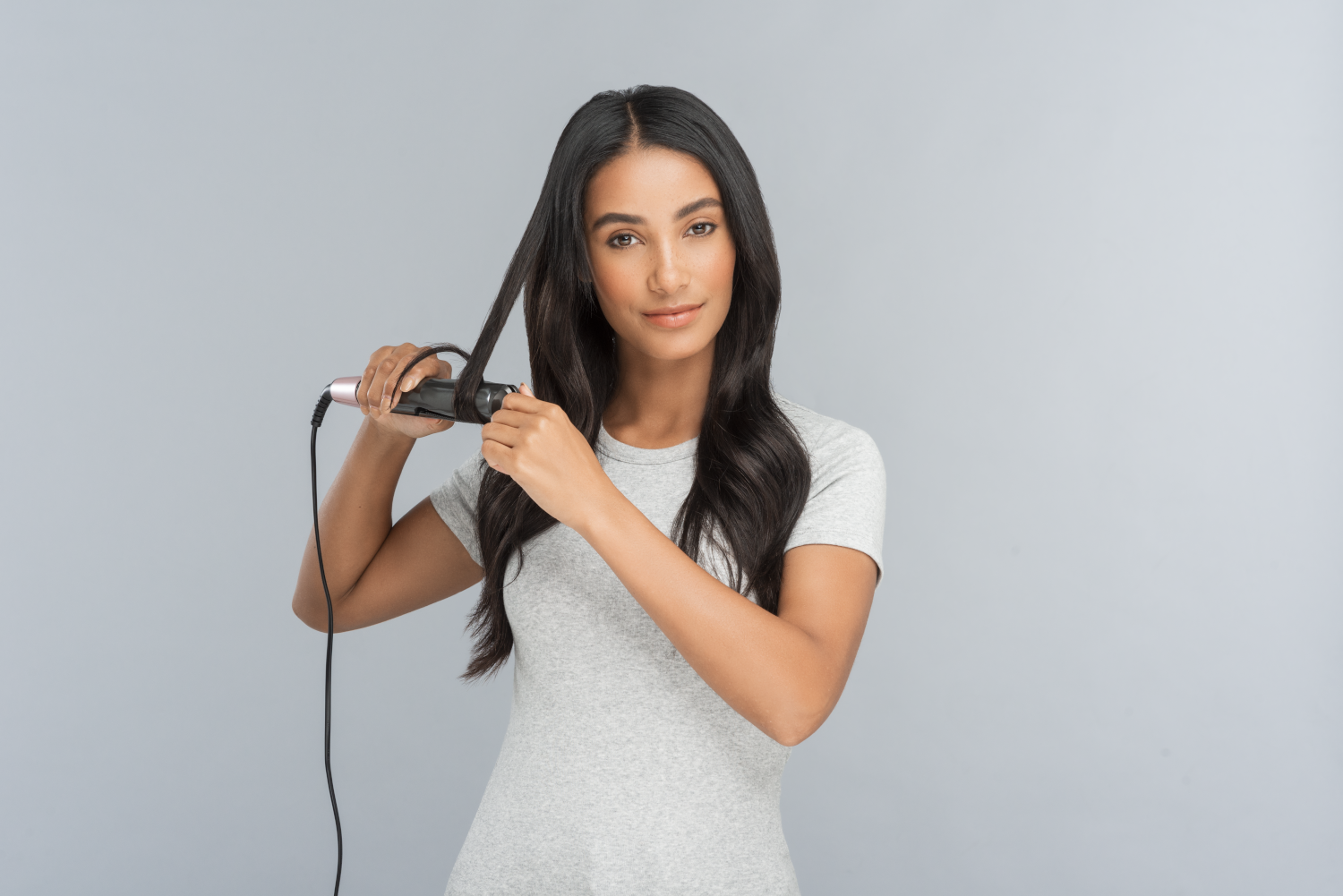 A woman in a gray shirt straightens her black hair with a curling iron, on gray background.