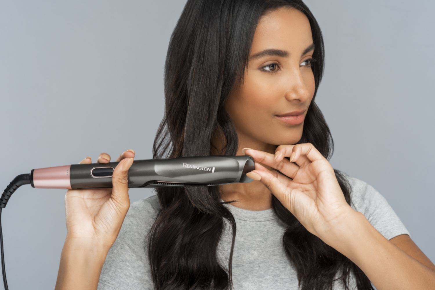 Woman using a hair straightener. She holds it near her hair, with a gray background.