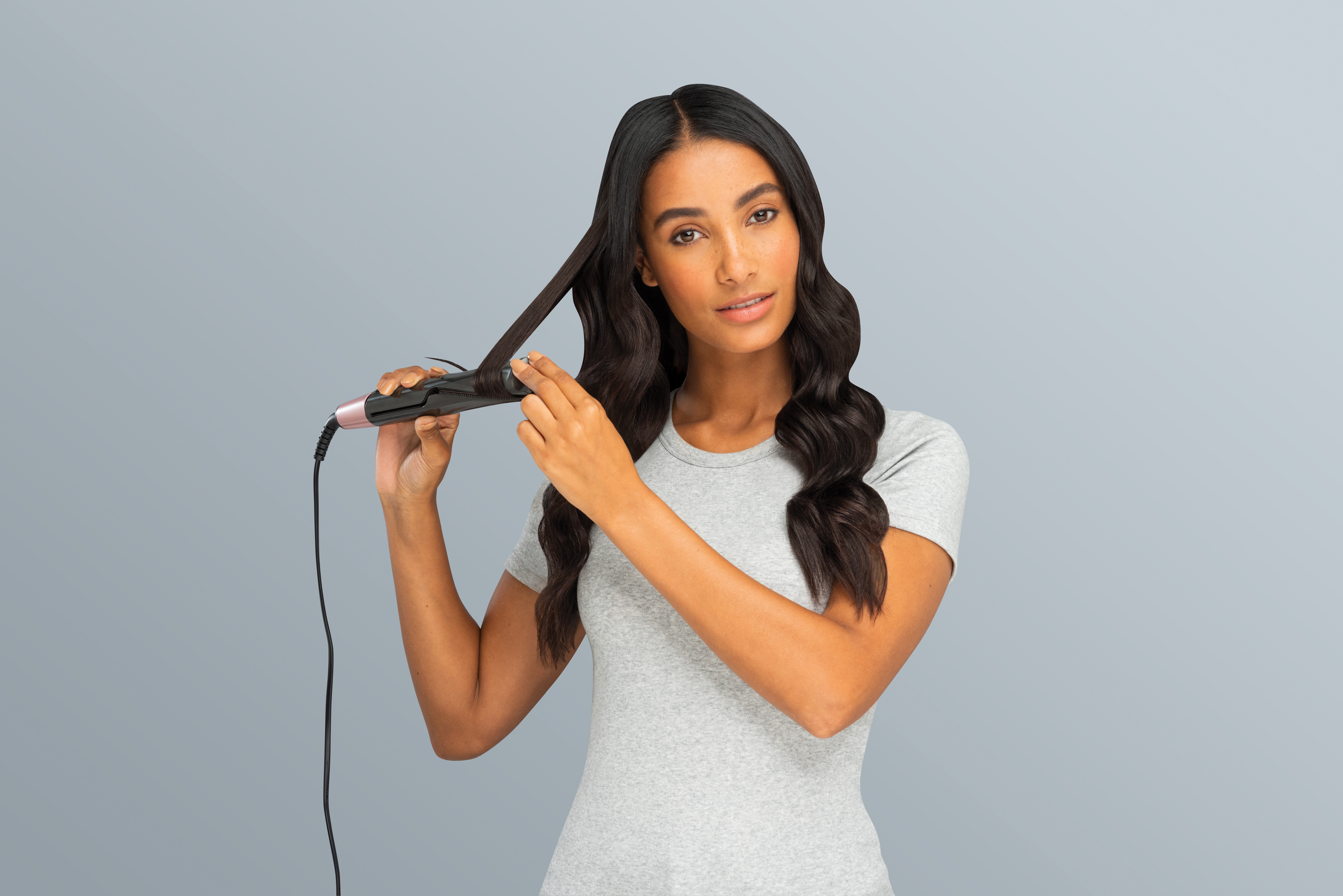 Woman curling hair with a flat iron. She wears a grey shirt, grey background.