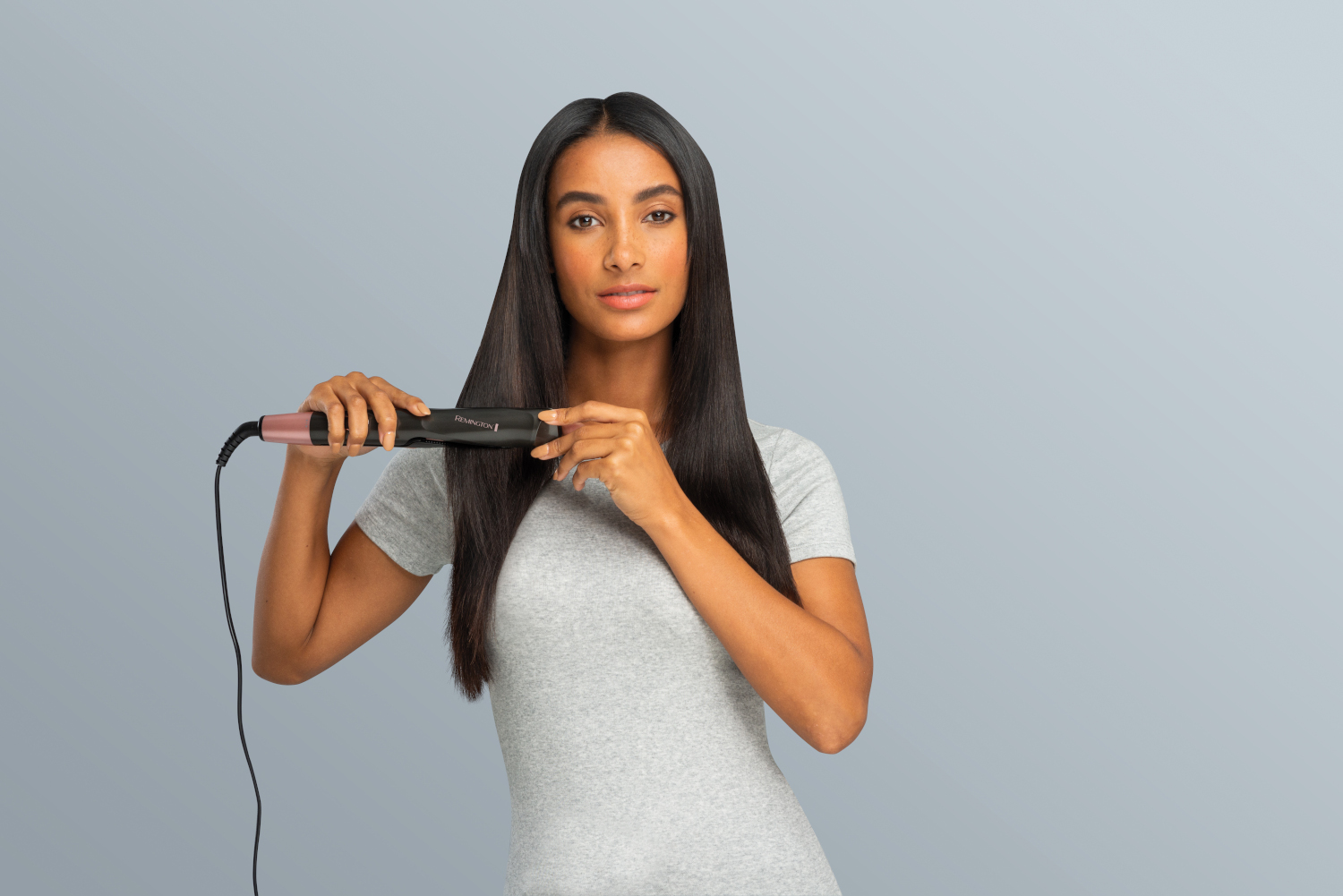 Woman using hair straightener. She holds the straightener in front of her straight black hair. Grey background.