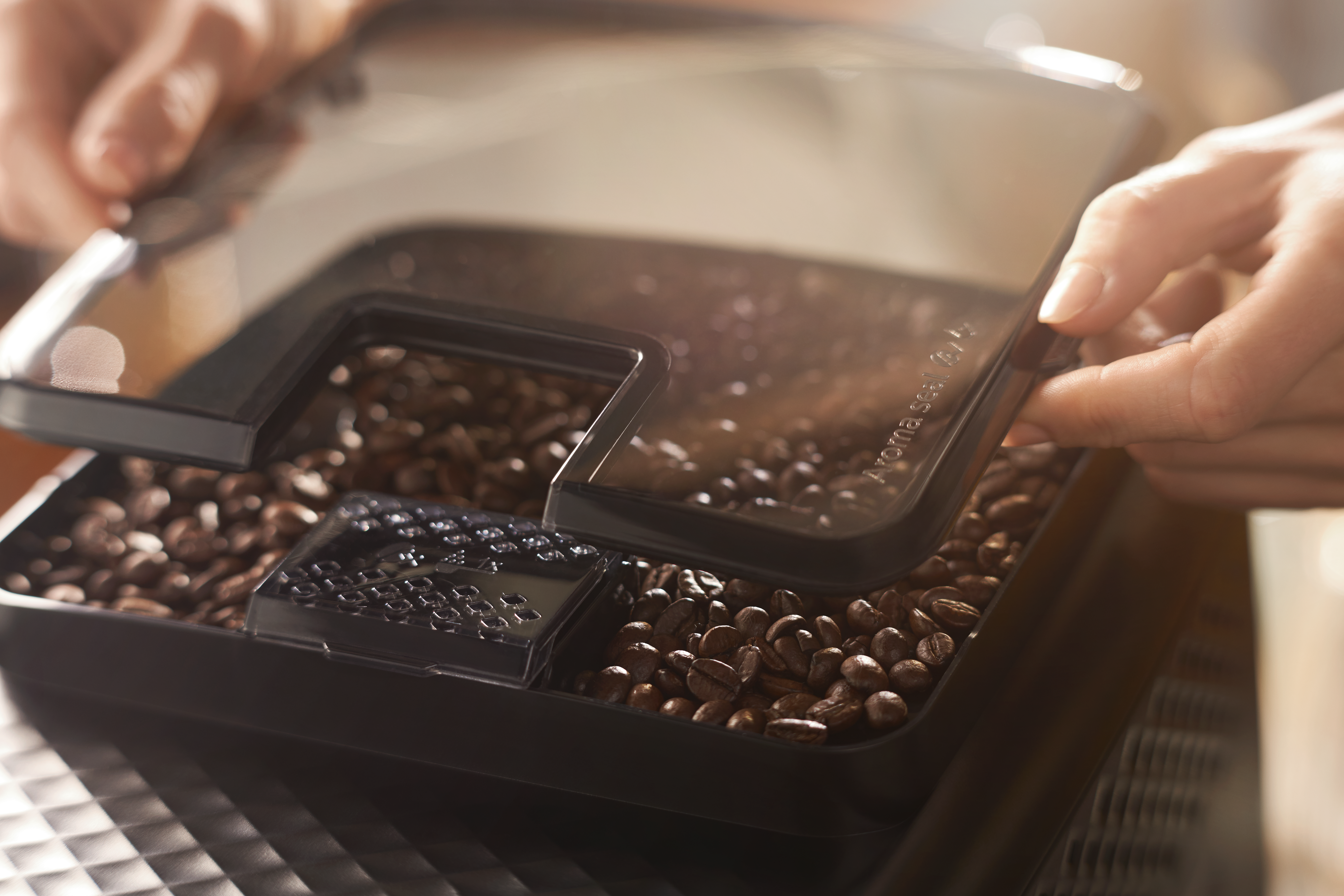 Una persona abre un recipiente de granos de café. El recipiente está lleno de granos de café. Se ven manos sosteniendo la tapa transparente.