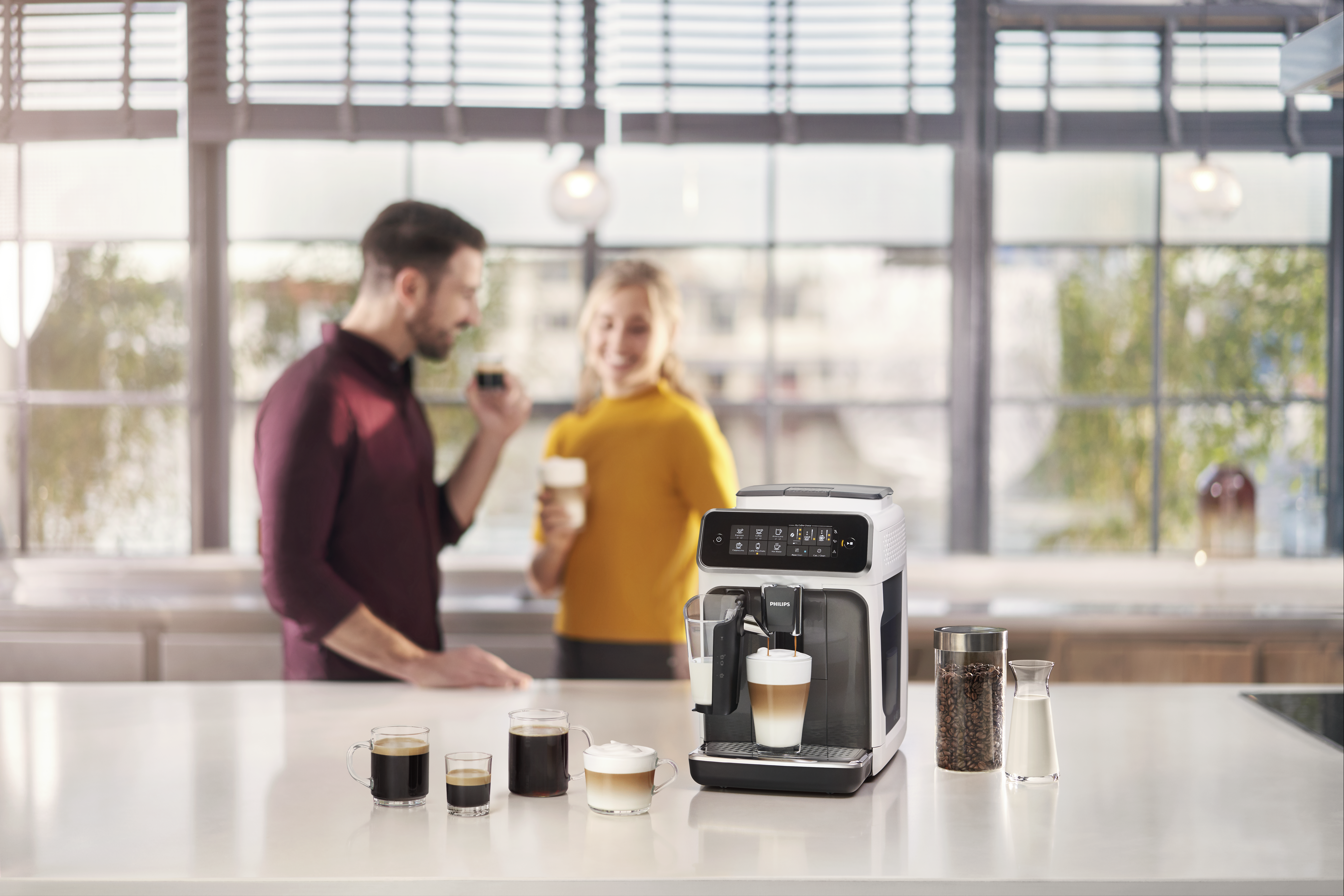 Una cafetera con tazas de café y dos personas en el fondo. Una sostiene una taza, la otra un vaso.