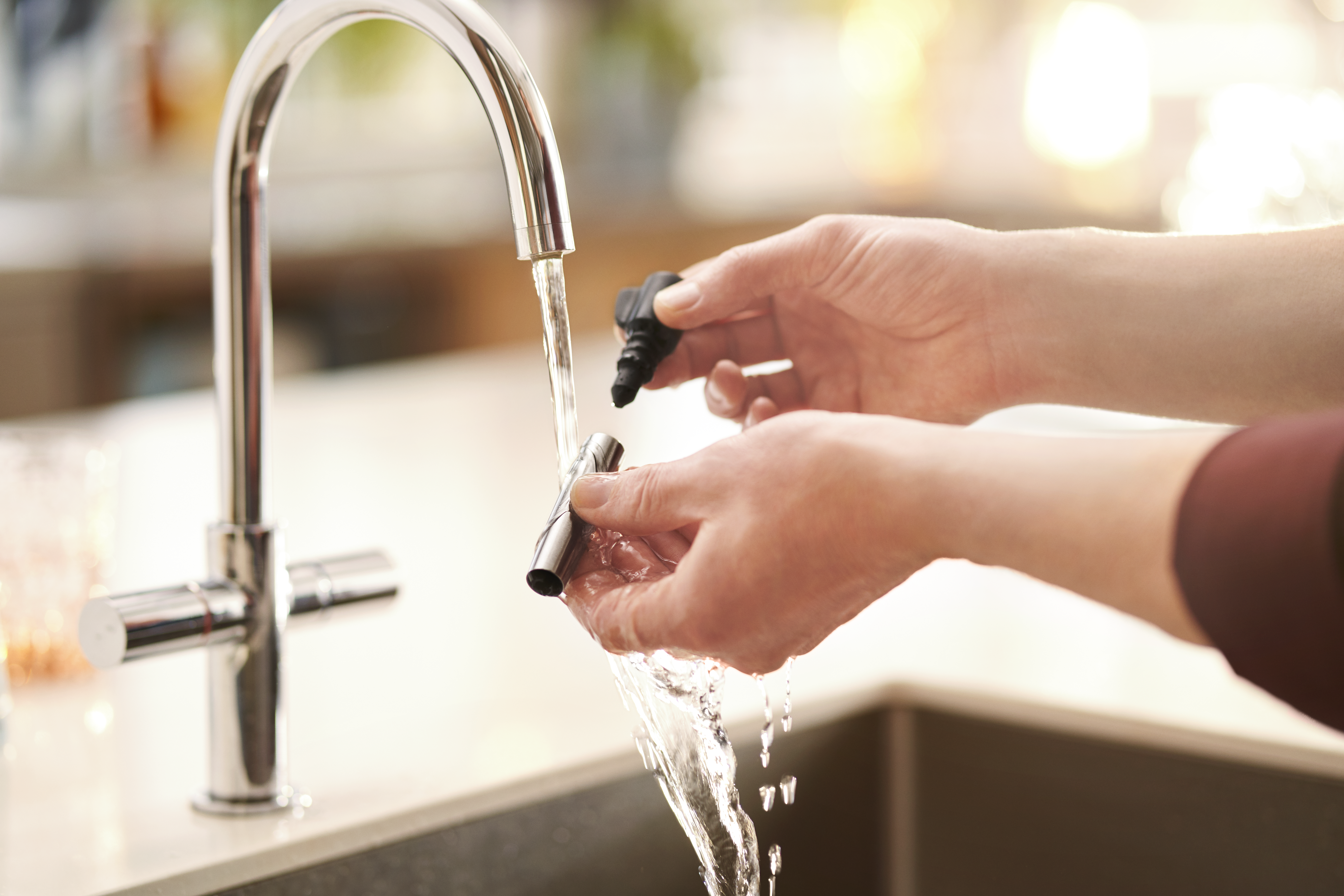 Manos sosteniendo un accesorio negro bajo agua corriente de un grifo cromado. La escena es interior, posiblemente una cocina.