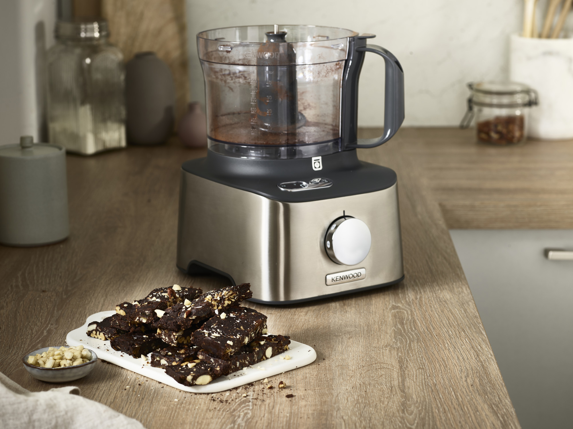 A food processor with brownies on a white plate. Ingredients and a wooden surface are also visible.