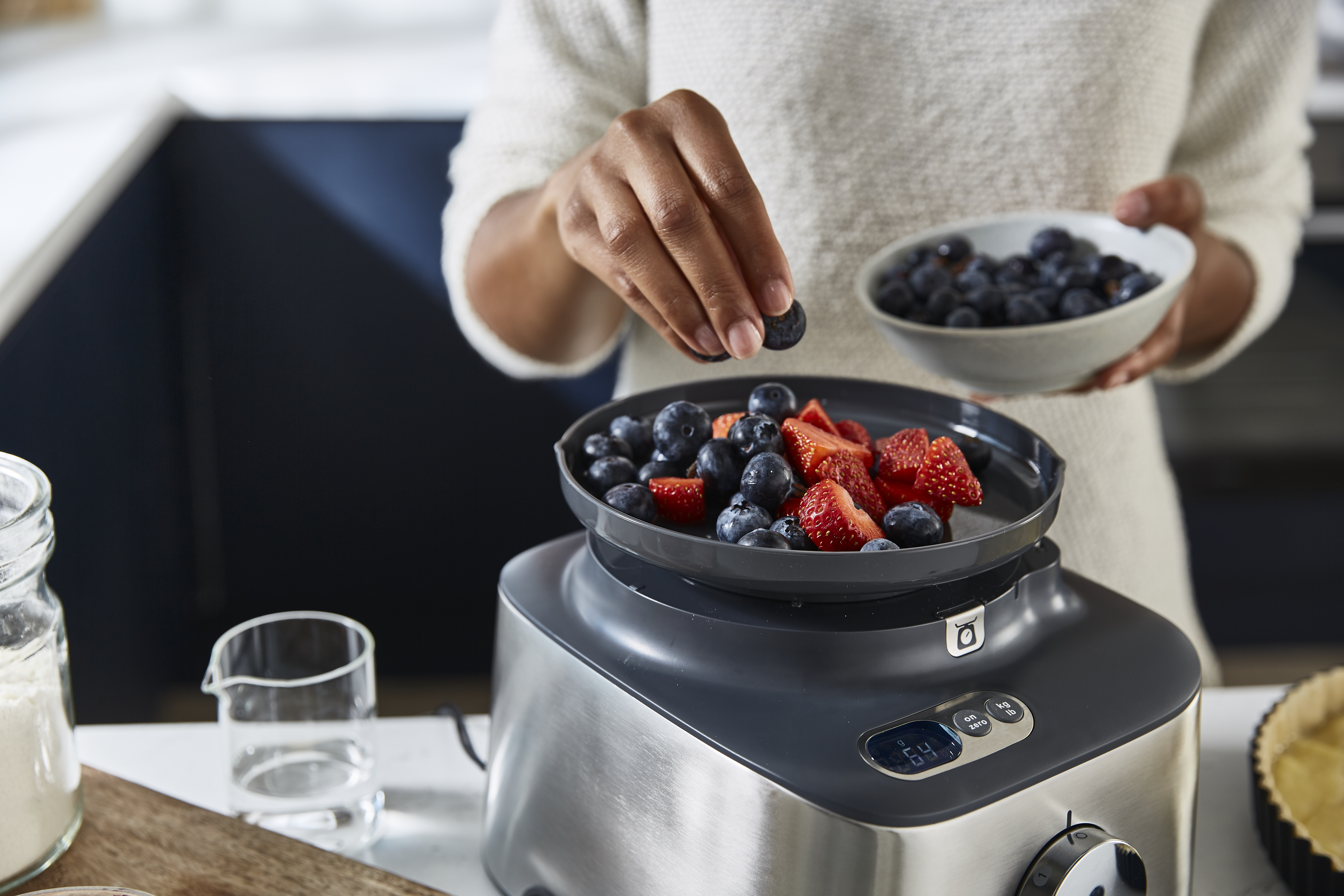 A person adds blueberries to a bowl of strawberries. A food processor is below.