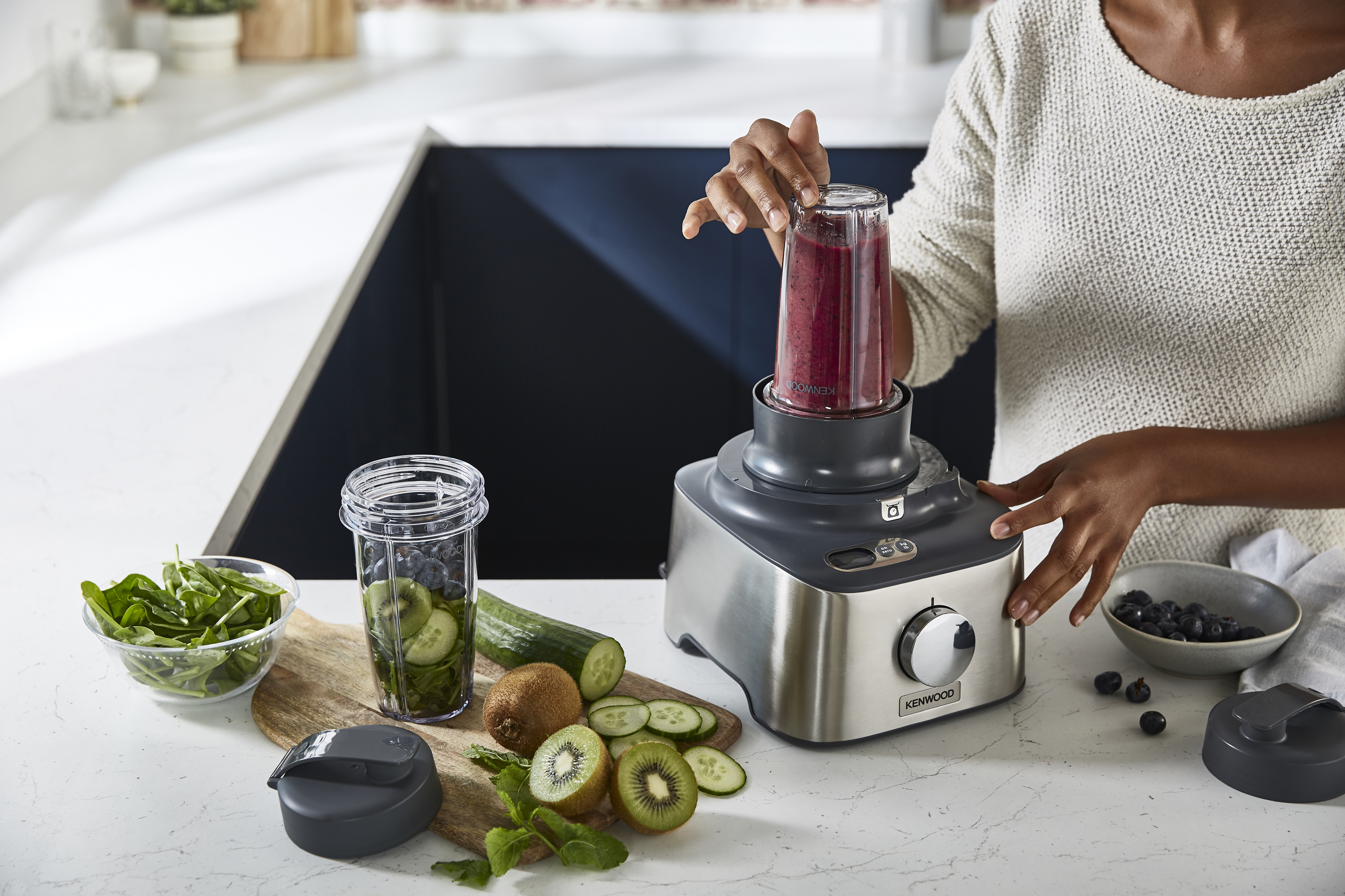 Woman using a blender to make smoothie, ingredients on the table. Kitchen setting with white counters.