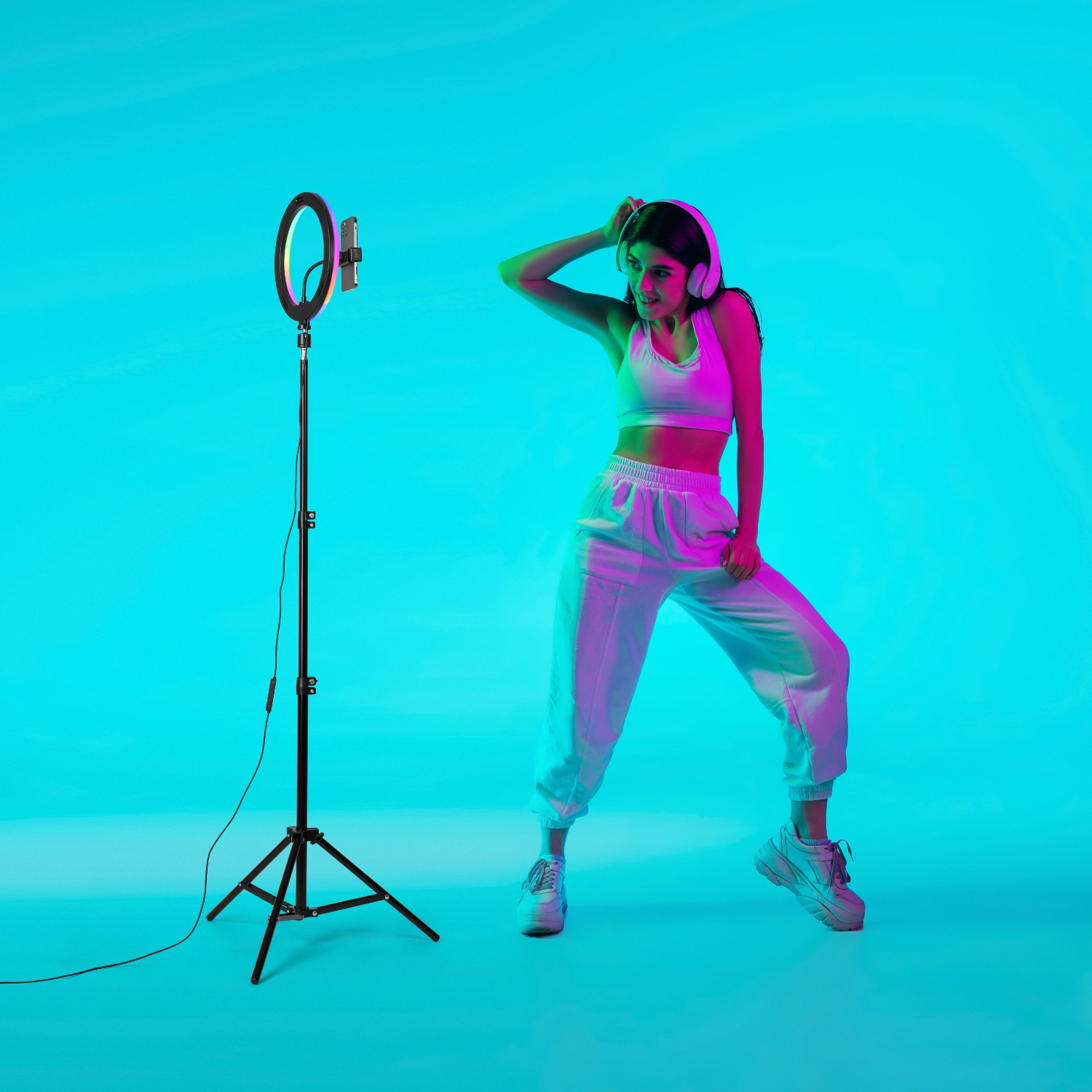 Woman wearing headphones and white outfit, standing by a phone attached to a ring light on a stand.